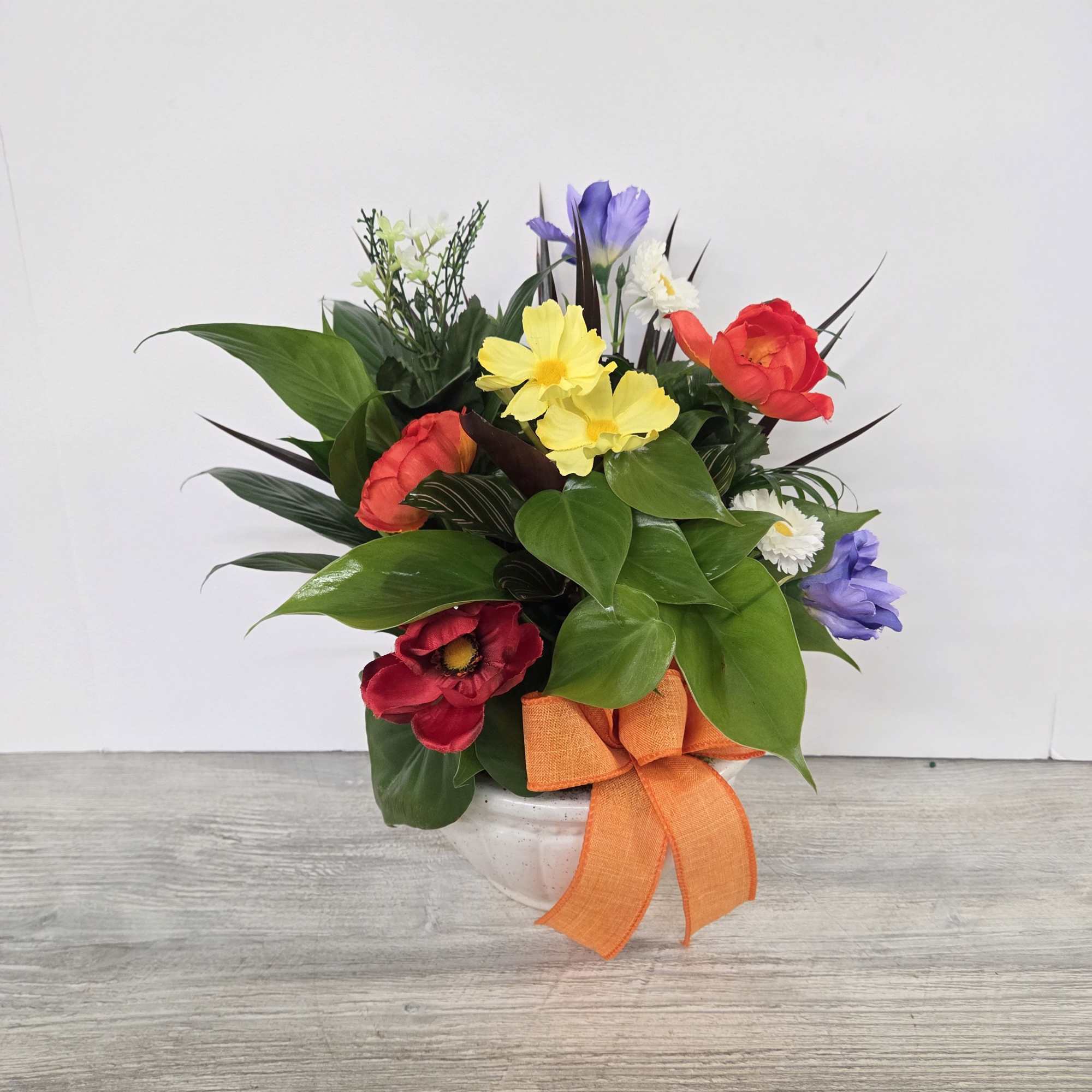 Colorful arrangement of red, yellow, and purple flowers in a white bowl with an orange bow.