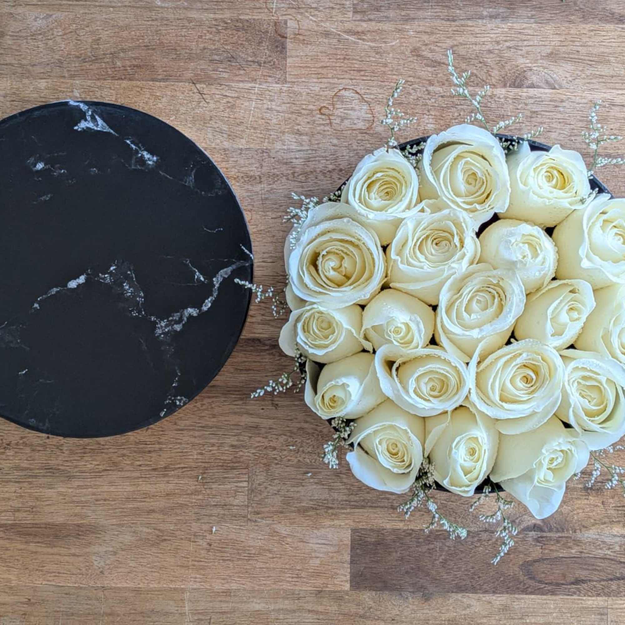 Round black box filled with cream roses, with a matching marble lid beside it on a wood surface.