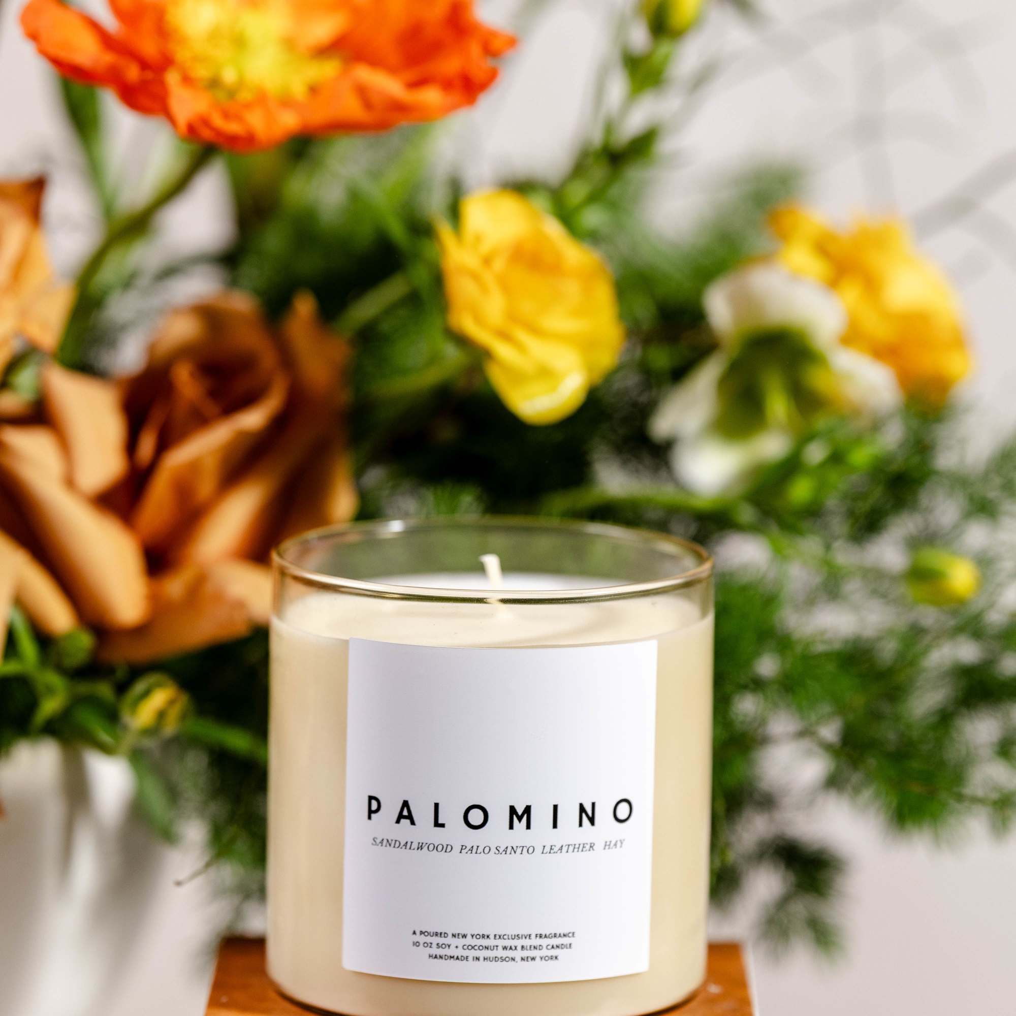 A scented candle in a glass jar sits in front of blurred orange and yellow flowers.