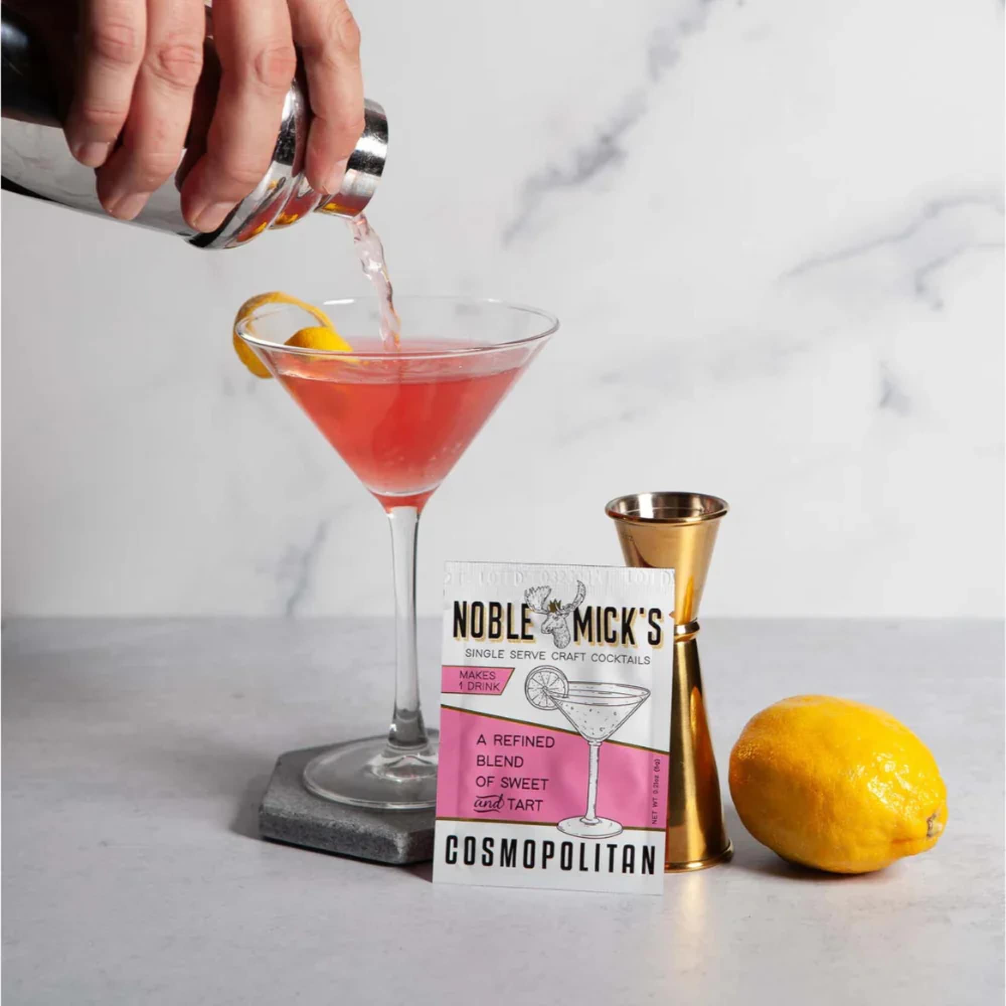 A hand pours a pink cocktail into a martini glass beside a mix packet and lemon.