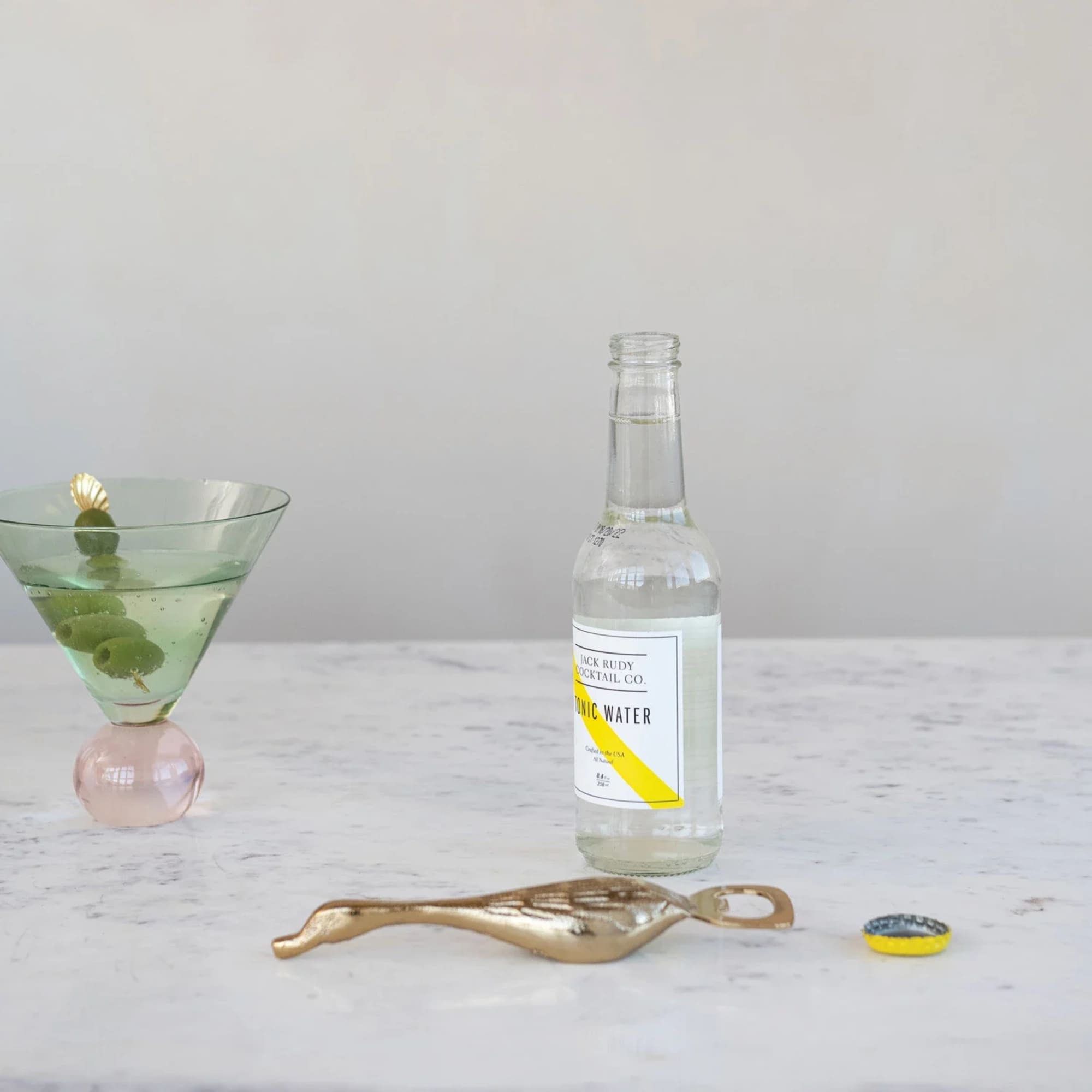 Green cocktail in a martini glass beside a tonic water bottle