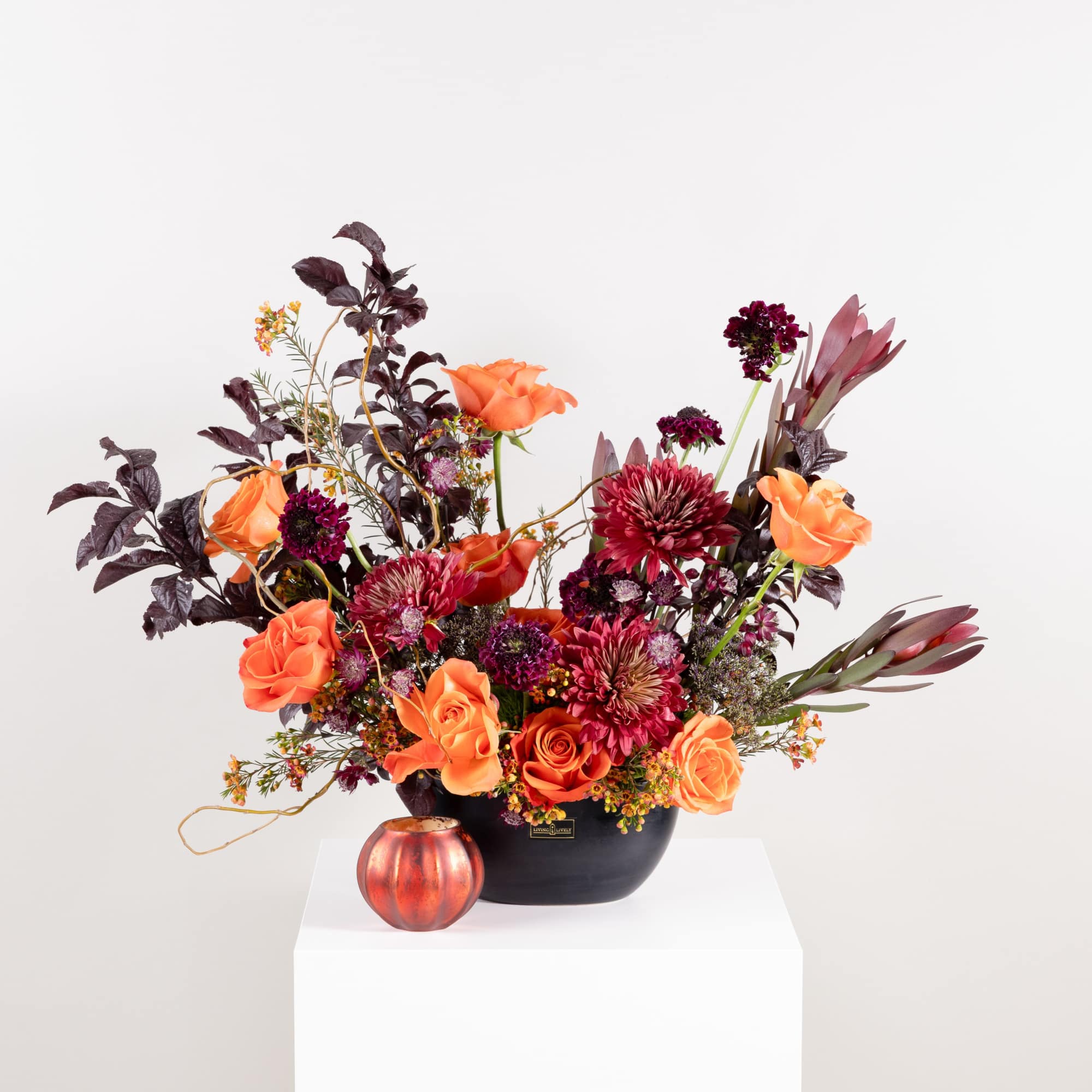 Orange and burgundy floral arrangement in a black vase with a small pumpkin candle