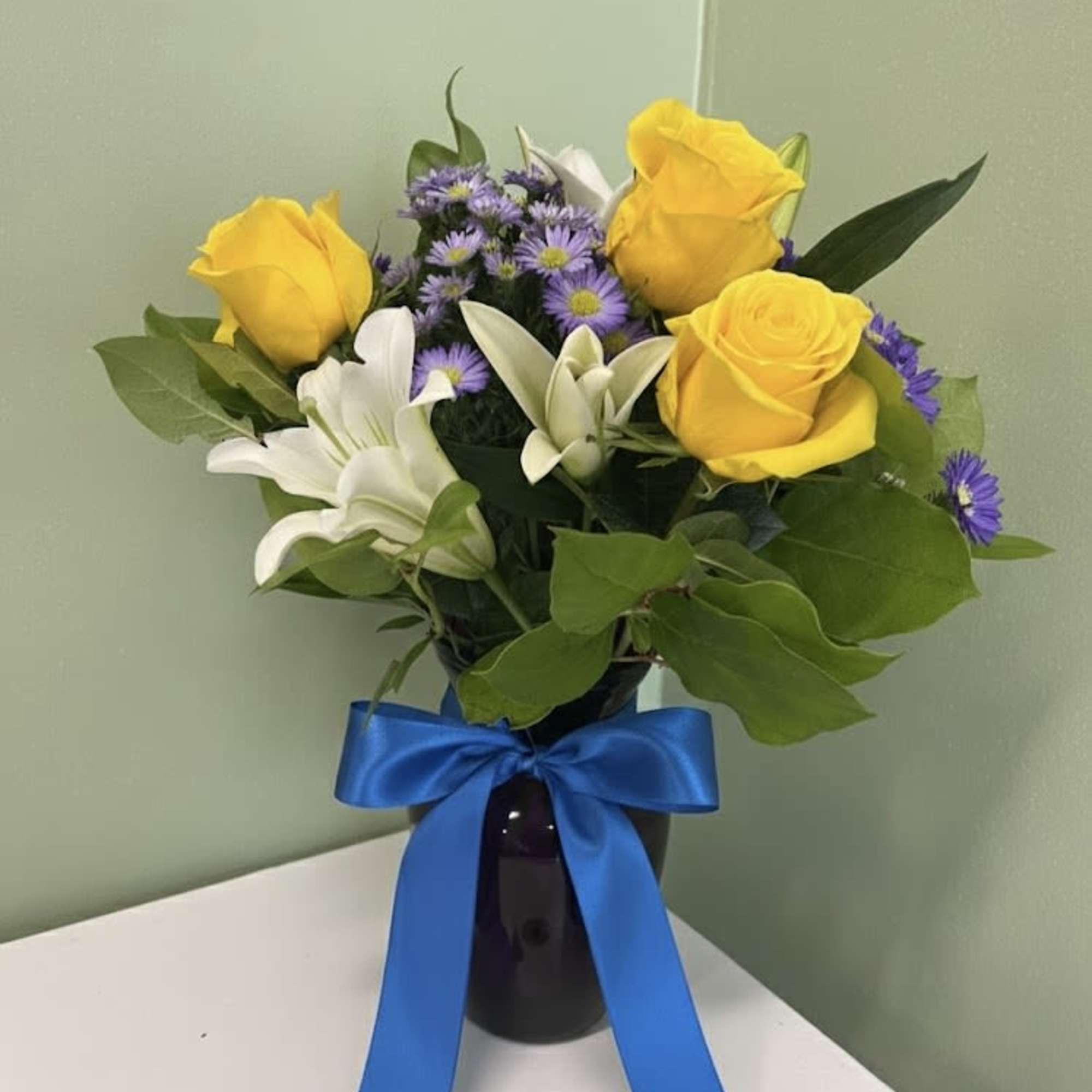 Arrangement of yellow roses, white lilies, and purple asters in a dark vase with a blue ribbon bow