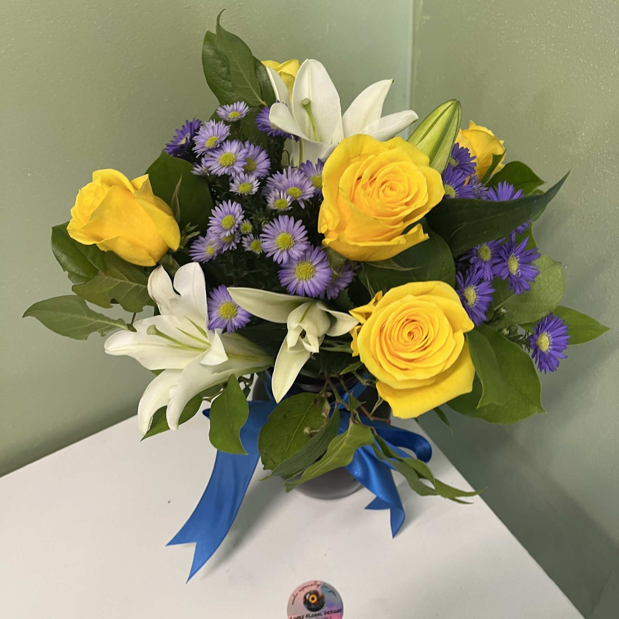 Everyday arrangement of yellow roses, white lilies, and purple asters with blue ribbon in a round container