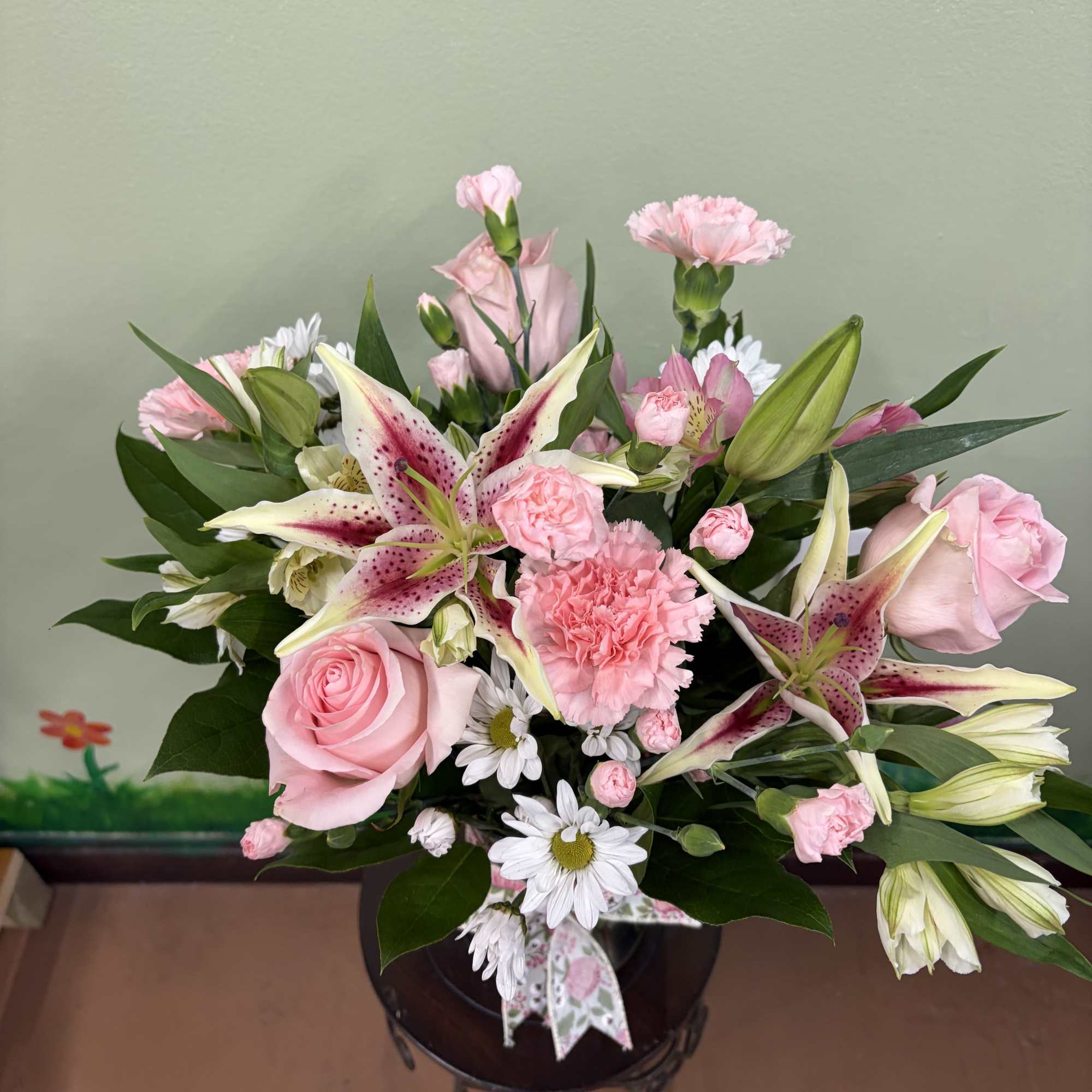 Pink lilies, roses, carnations, and white daisies arranged in a clear glass vase with a bow.