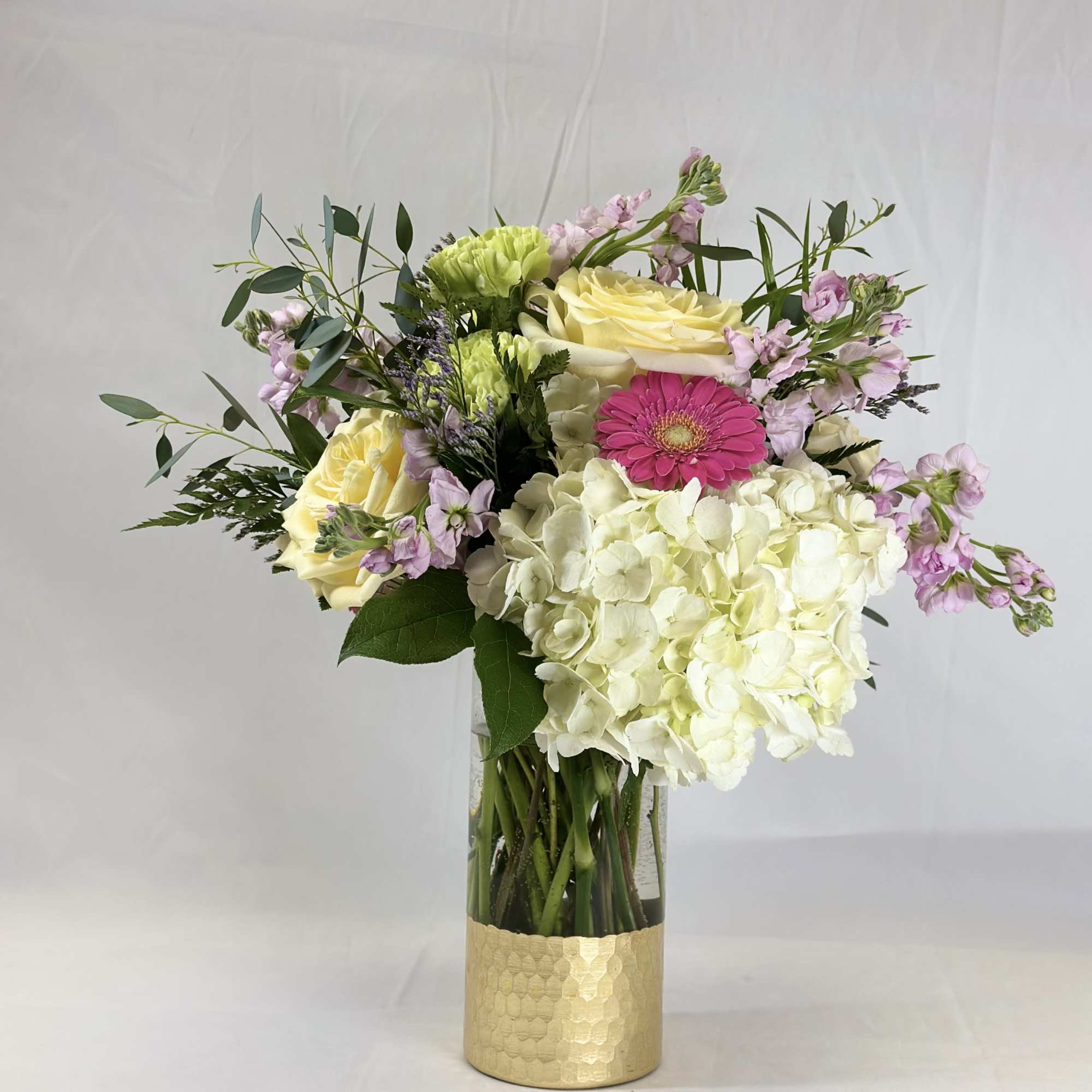 Mixed bouquet of white hydrangeas, pale yellow roses, and pink gerbera daisies in a gold vase