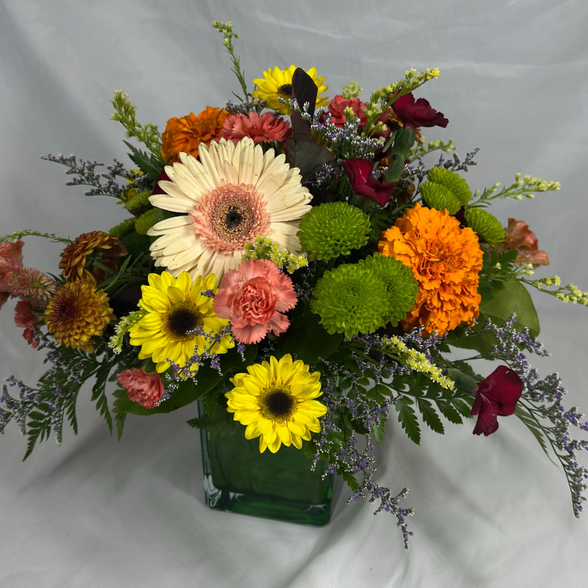 Mixed bouquet of sunflowers, gerbera daisies, and carnations in a green glass vase