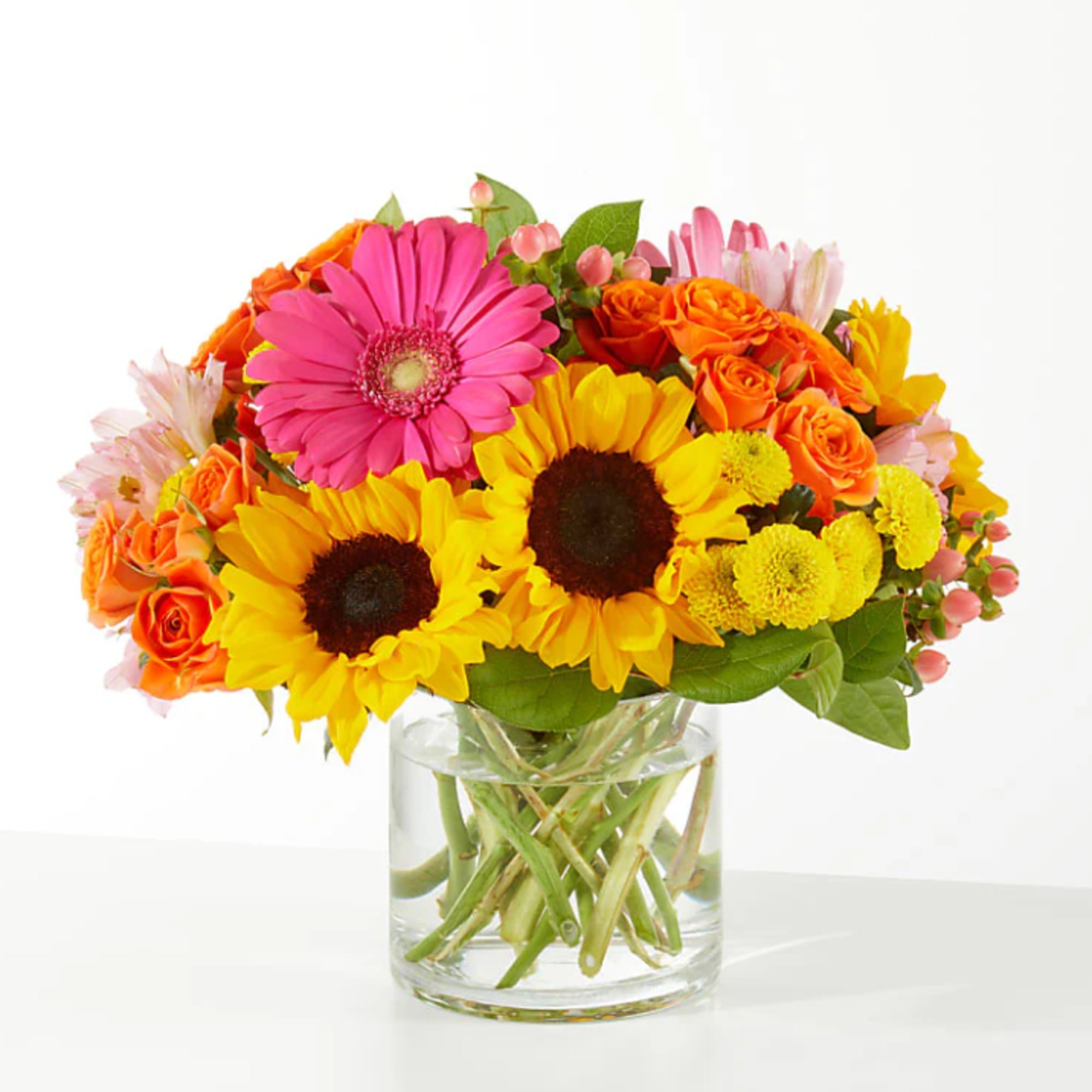 Bright mixed bouquet in a clear glass vase with sunflowers, roses, and a pink gerbera daisy