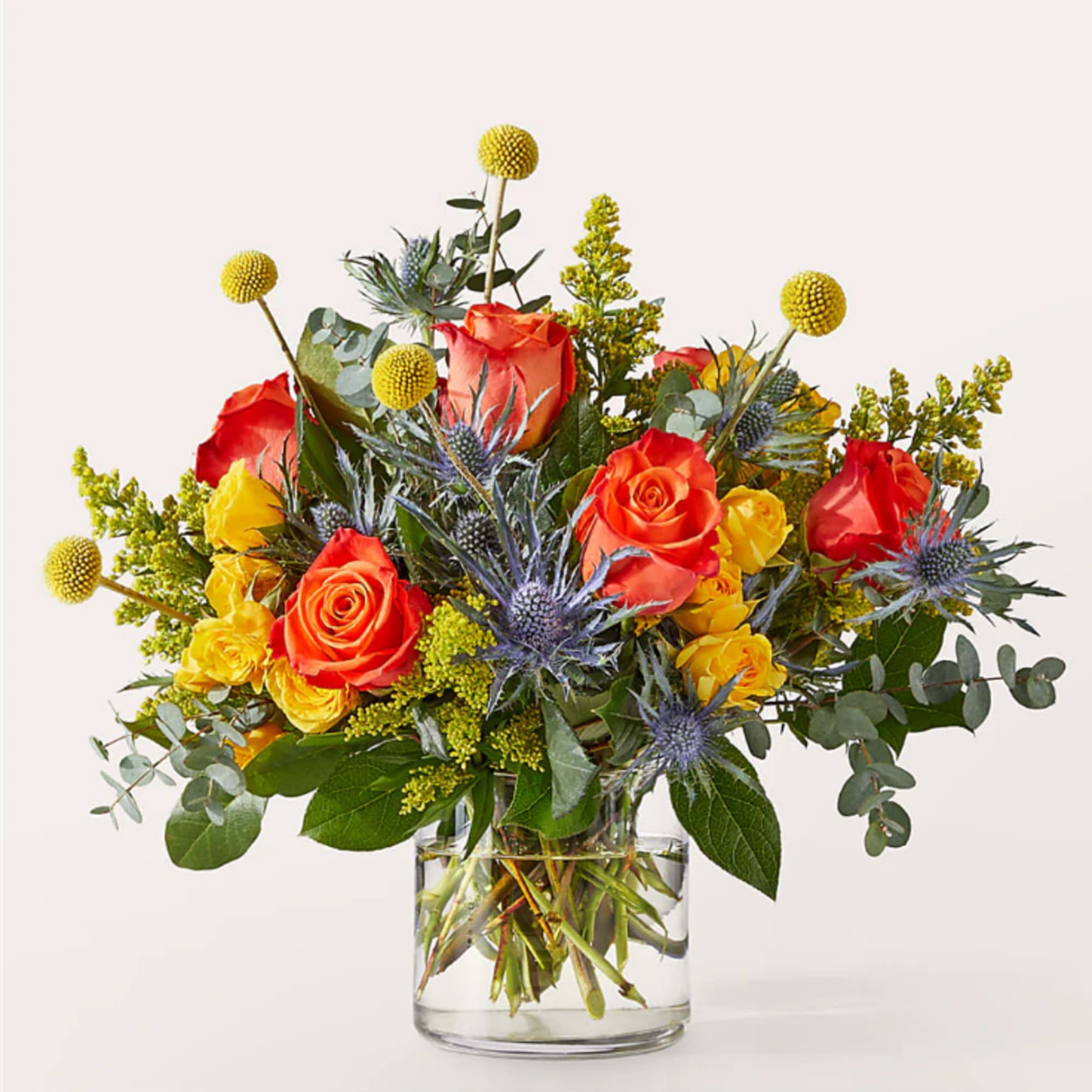 Orange and yellow roses in a clear glass vase with blue thistle accents