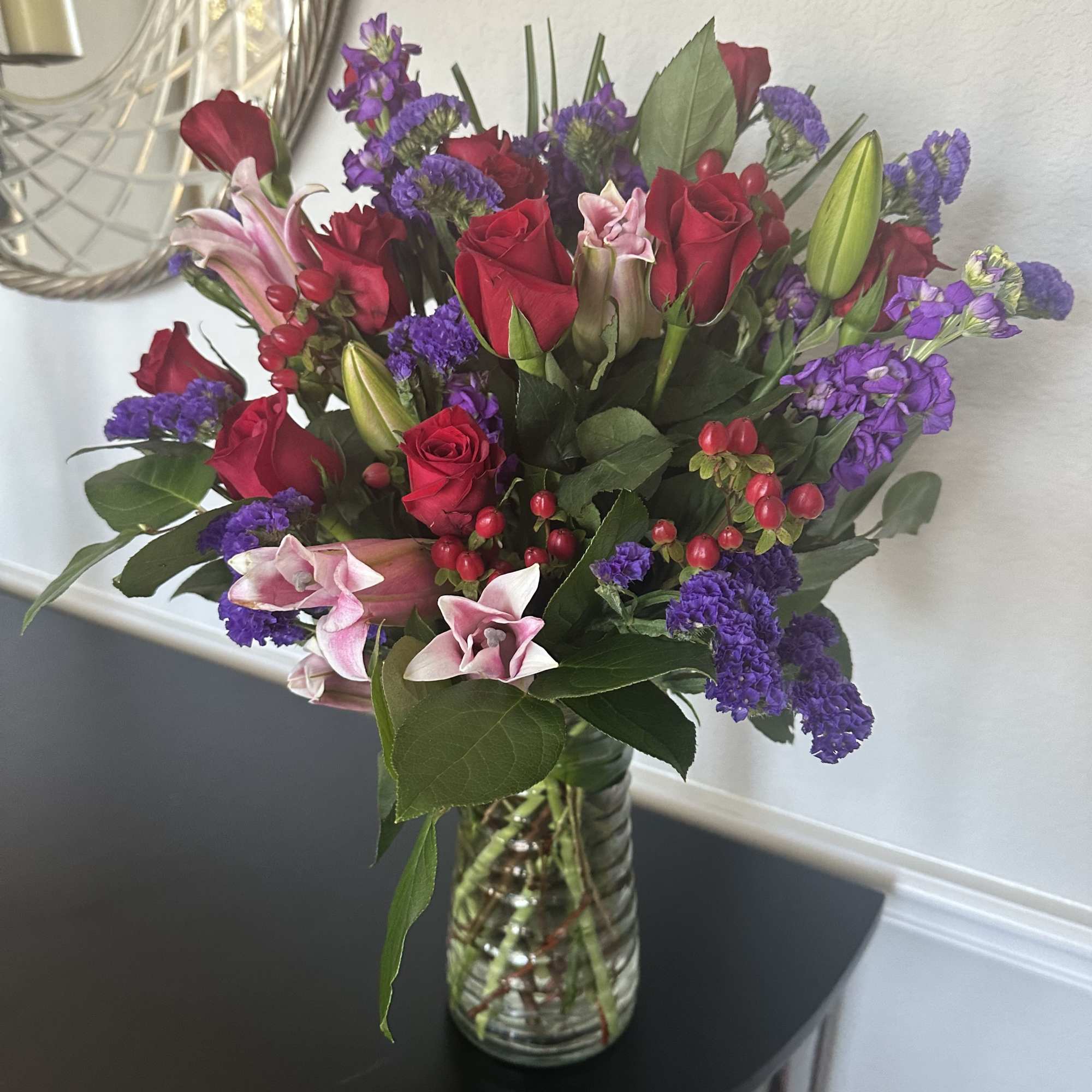 Mixed bouquet of red roses, pink lilies, and purple flowers arranged in a clear glass vase.