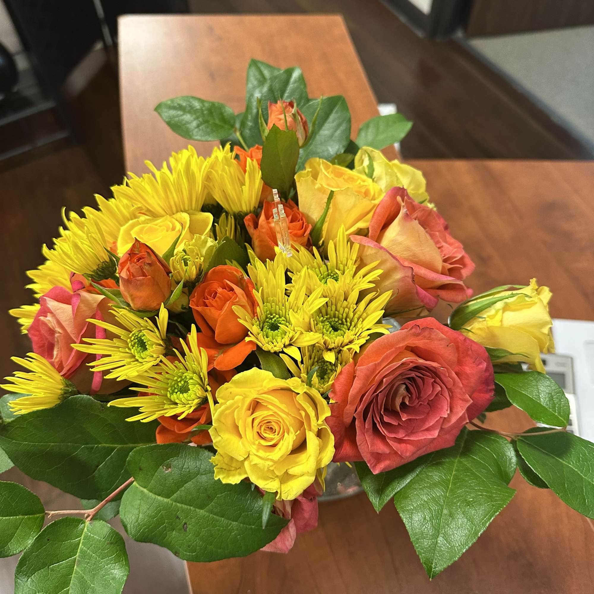 Low arrangement of yellow and orange roses and yellow mums in a clear glass vase on a wooden table.