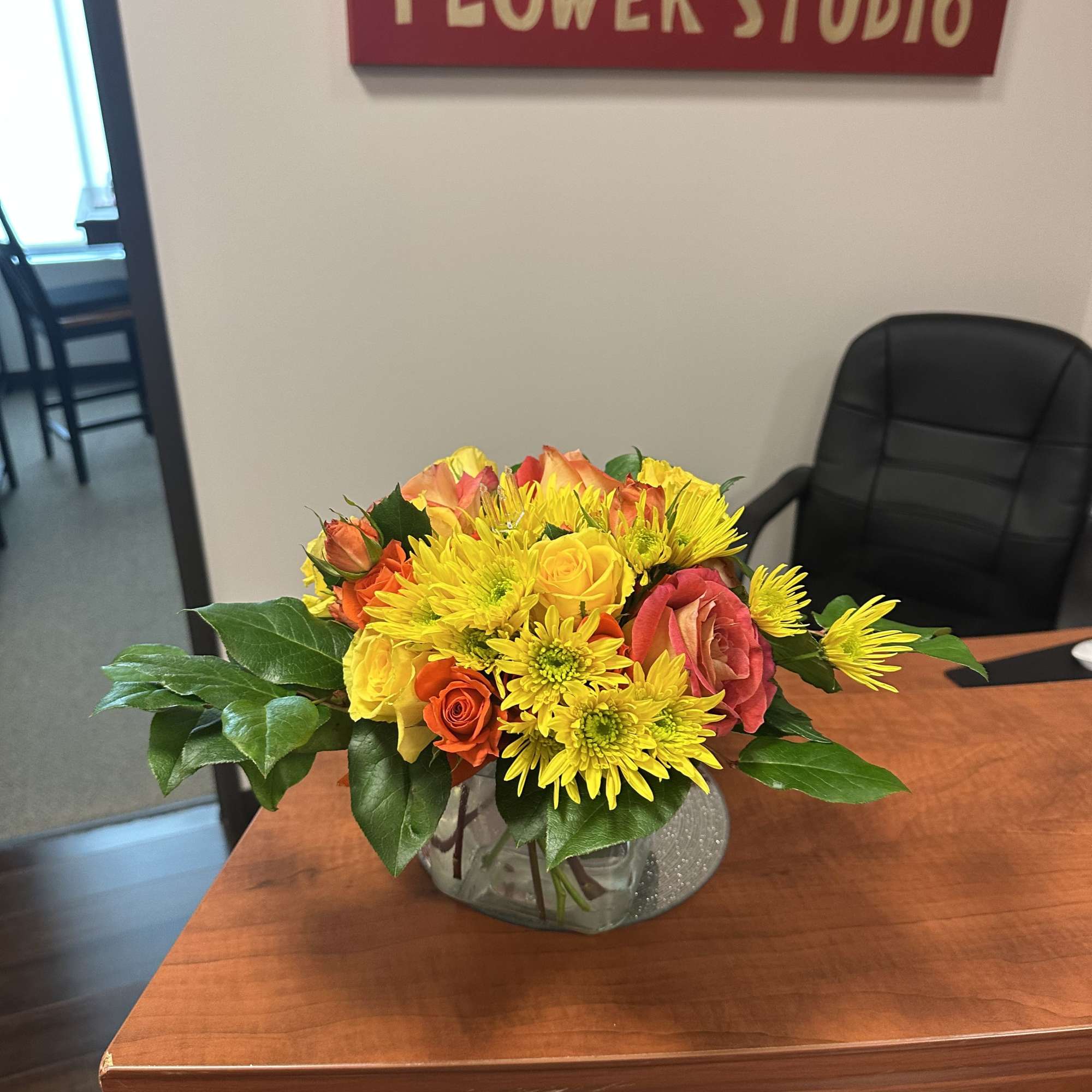 Low arrangement of yellow mums and orange and yellow roses in a clear glass vase on a desk