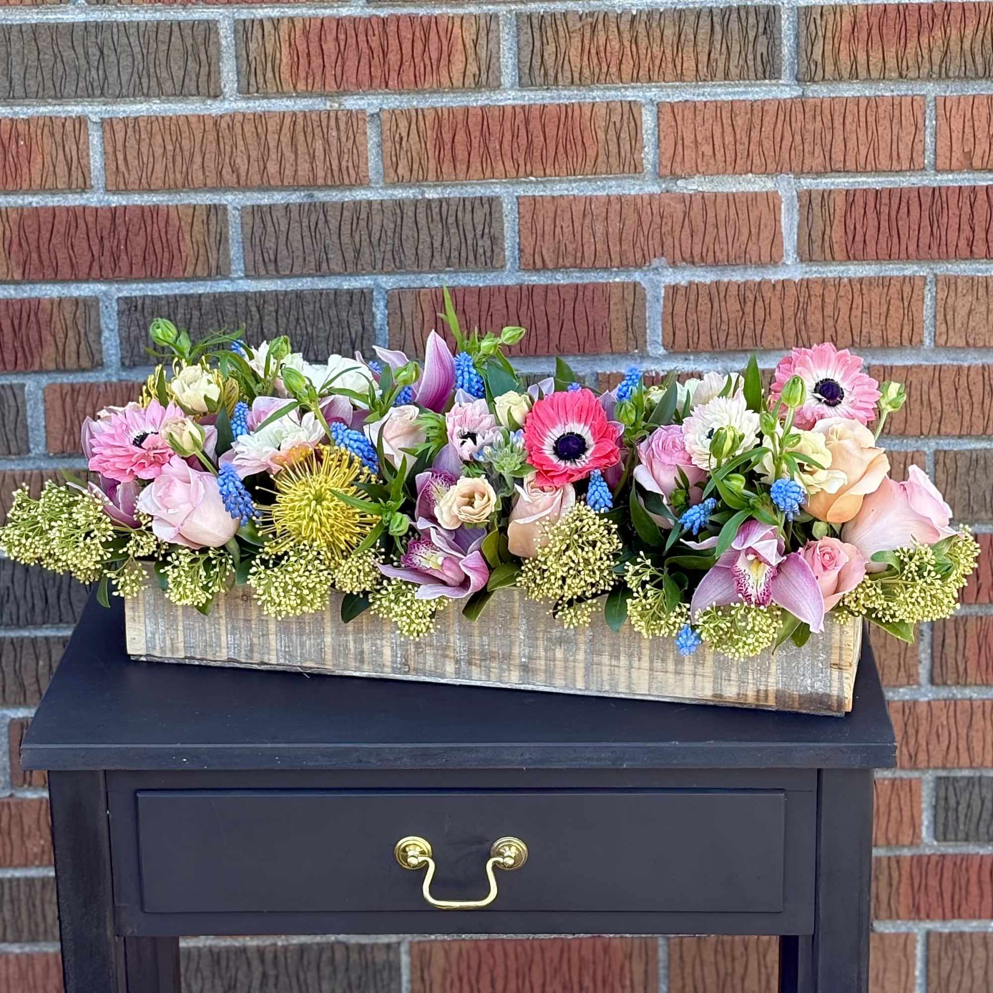 Low pastel flower arrangement in a rustic wooden box on a black table against a brick wall