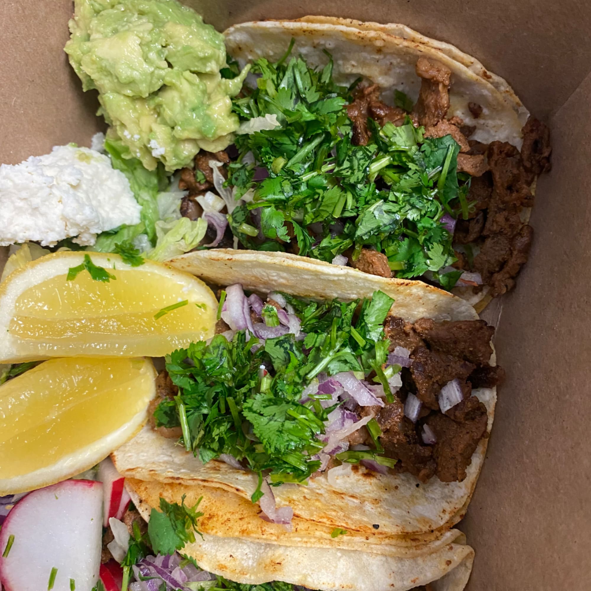 Two beef tacos with cilantro, onion, lime wedges, and guacamole in a takeout box