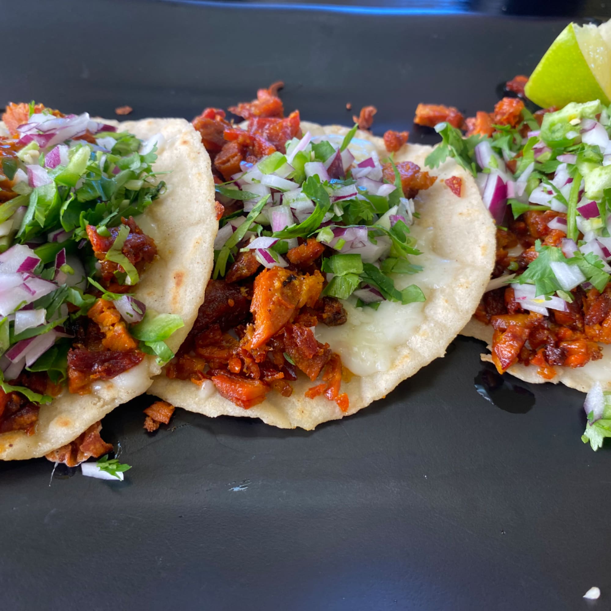 Three tacos topped with chopped onions, cilantro, and lime on a black plate