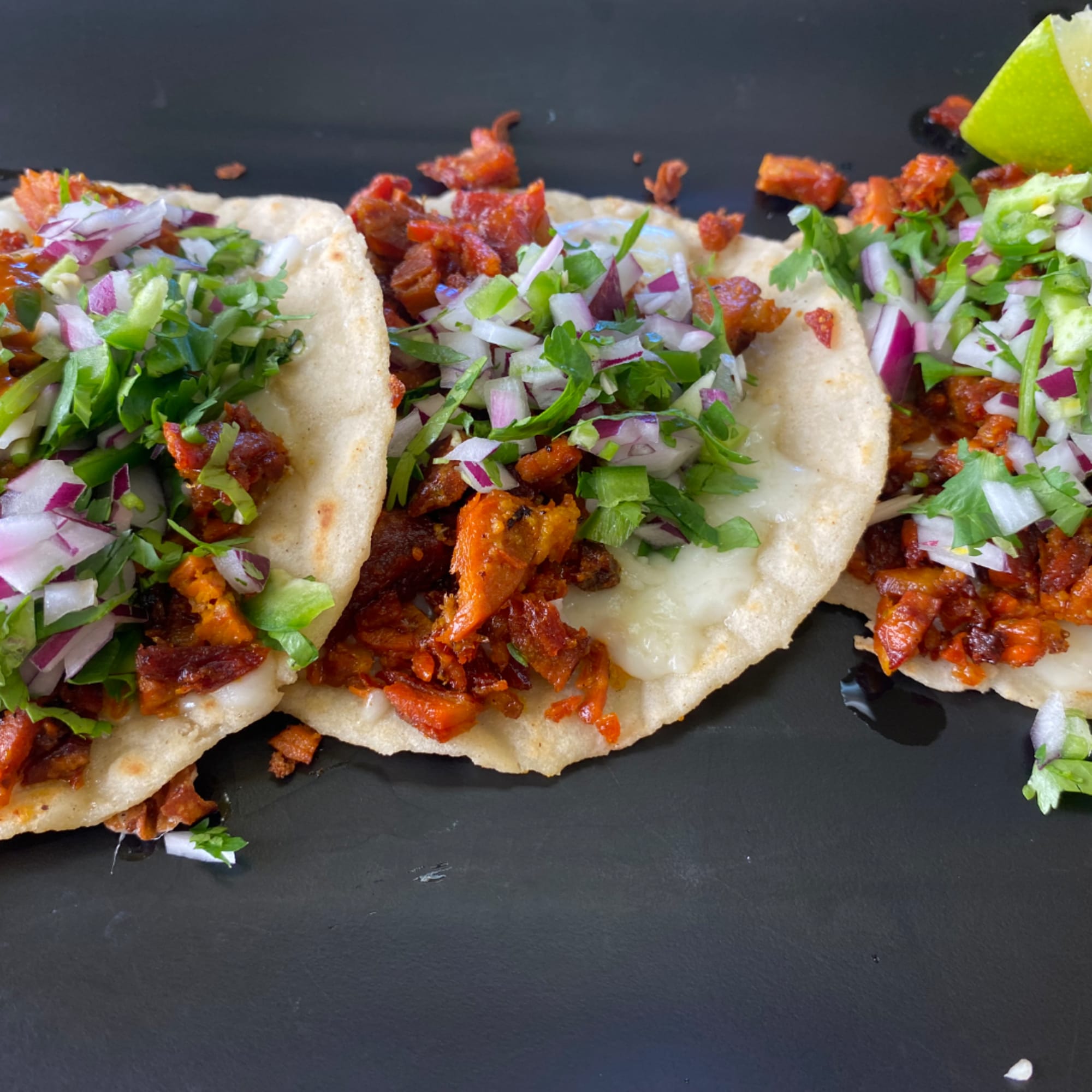 Three tacos topped with chopped onions, cilantro, and meat on a black tray