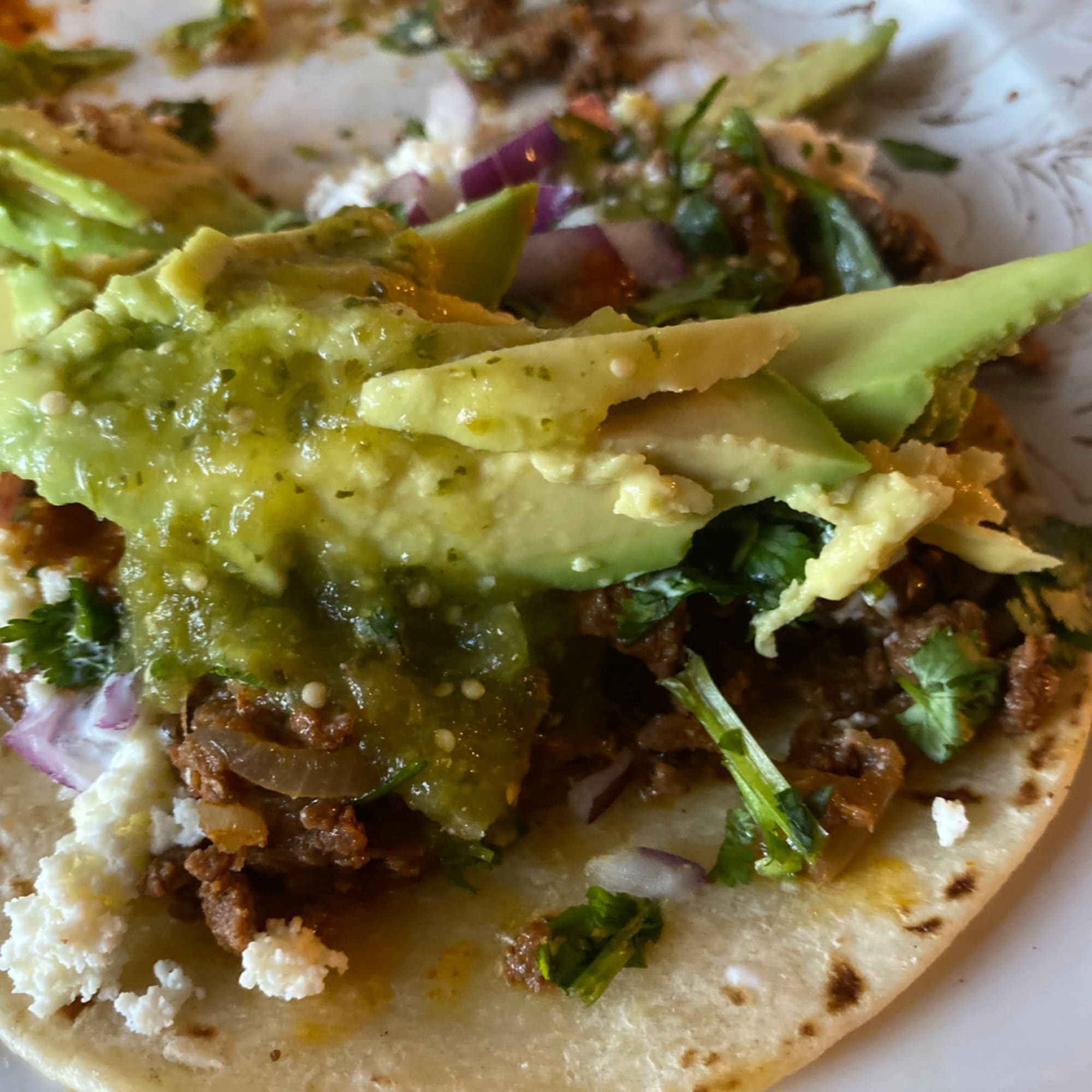 Tacos topped with avocado slices, green salsa, and crumbled cheese on a plate.