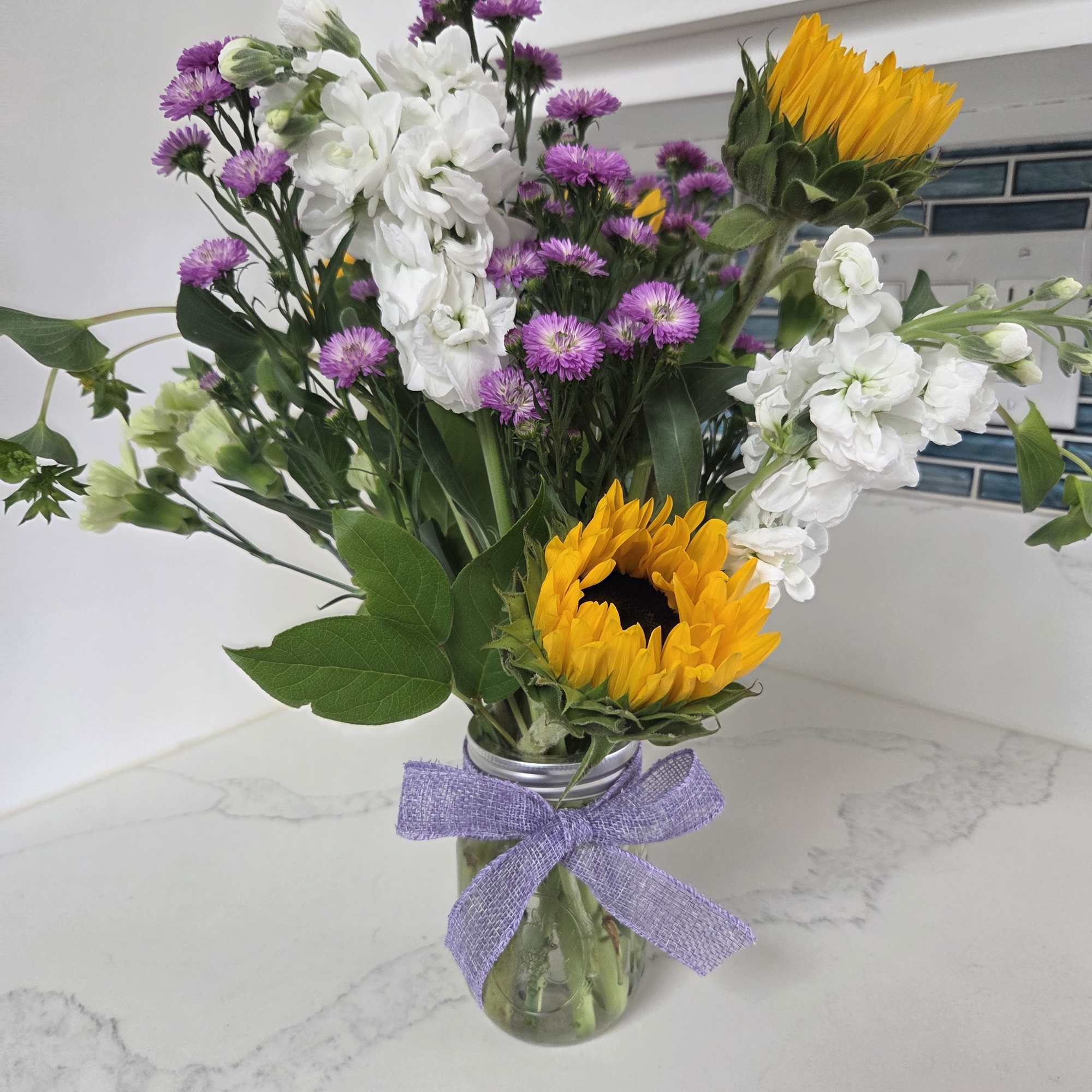 Bouquet of sunflowers, white flowers, and purple blooms in a glass jar
