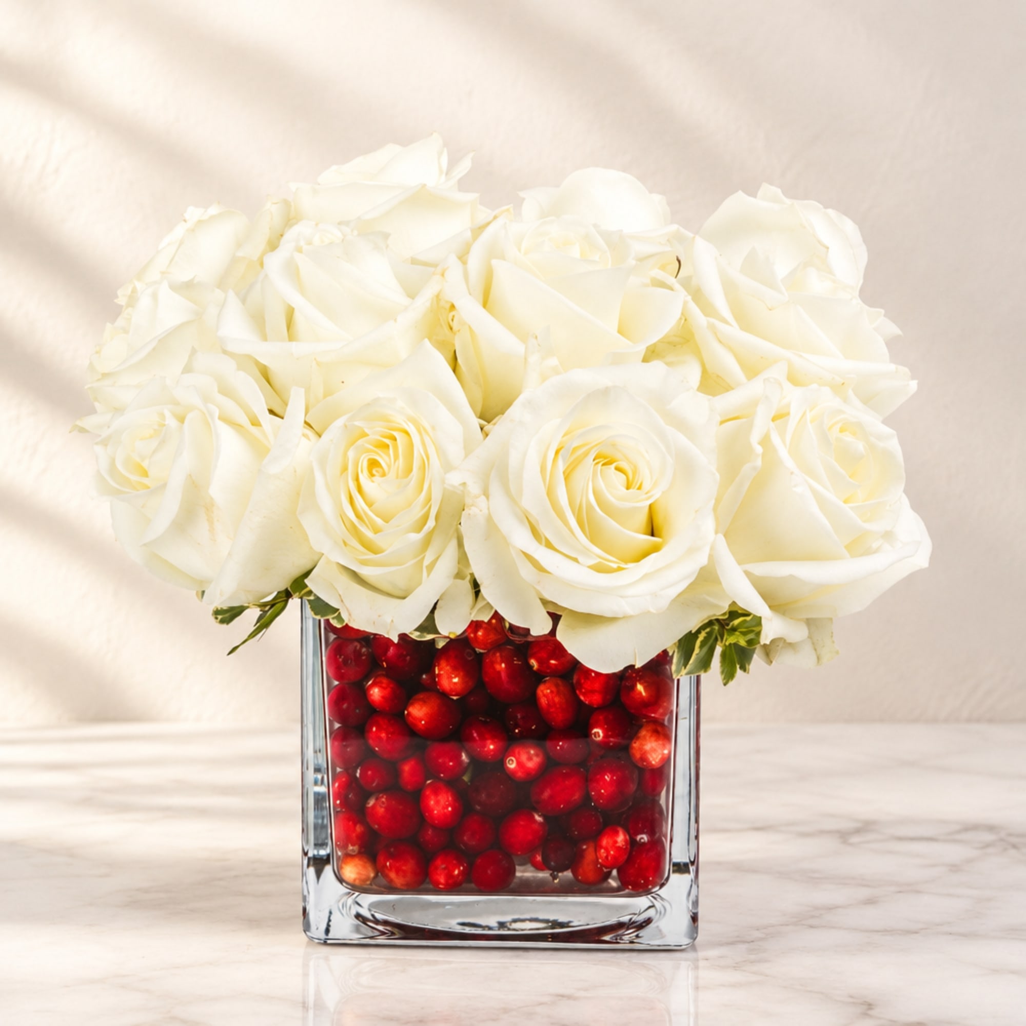 White roses arranged in a square glass vase filled with red berries
