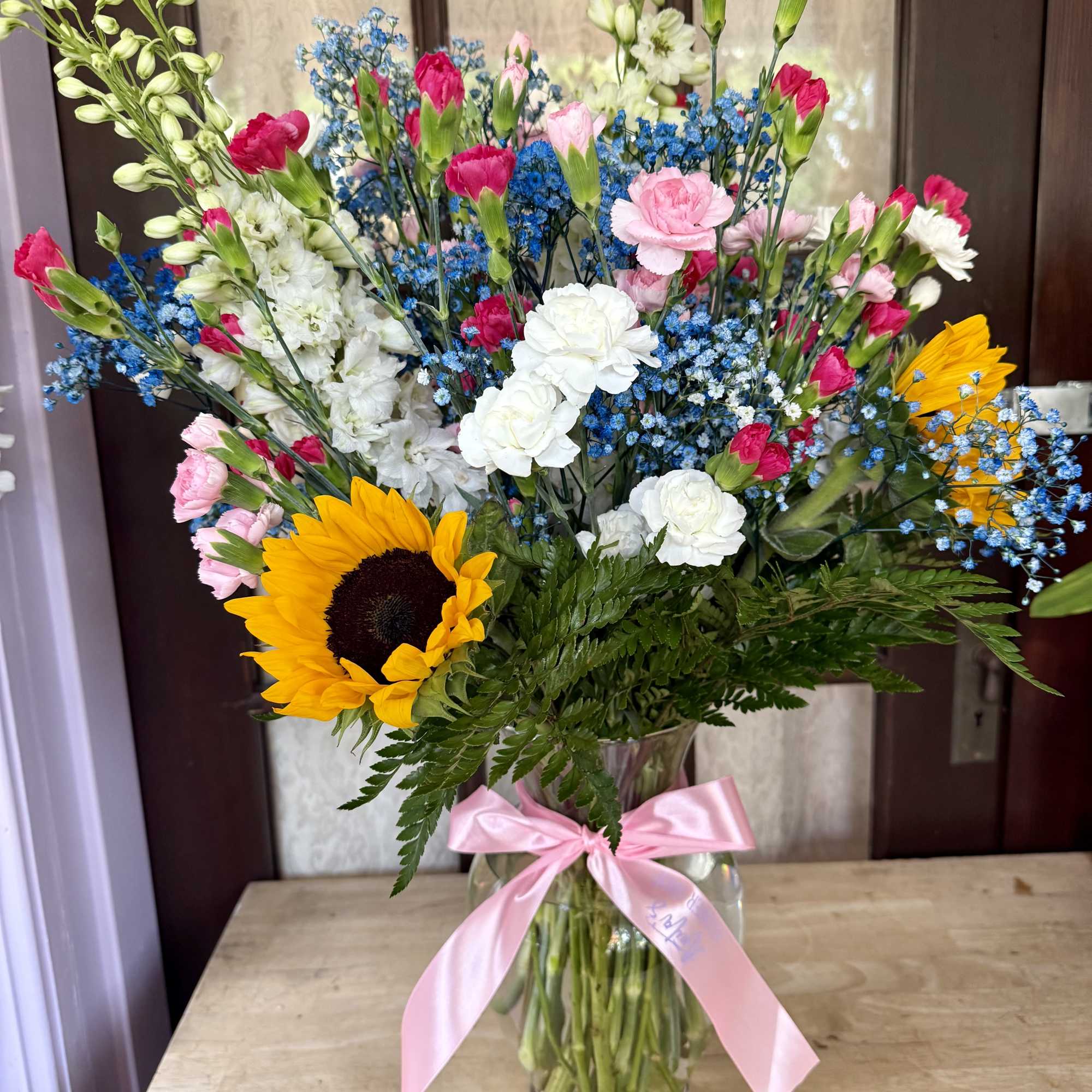 Mixed bouquet with sunflowers, carnations, and blue filler in a glass vase with a pink ribbon.