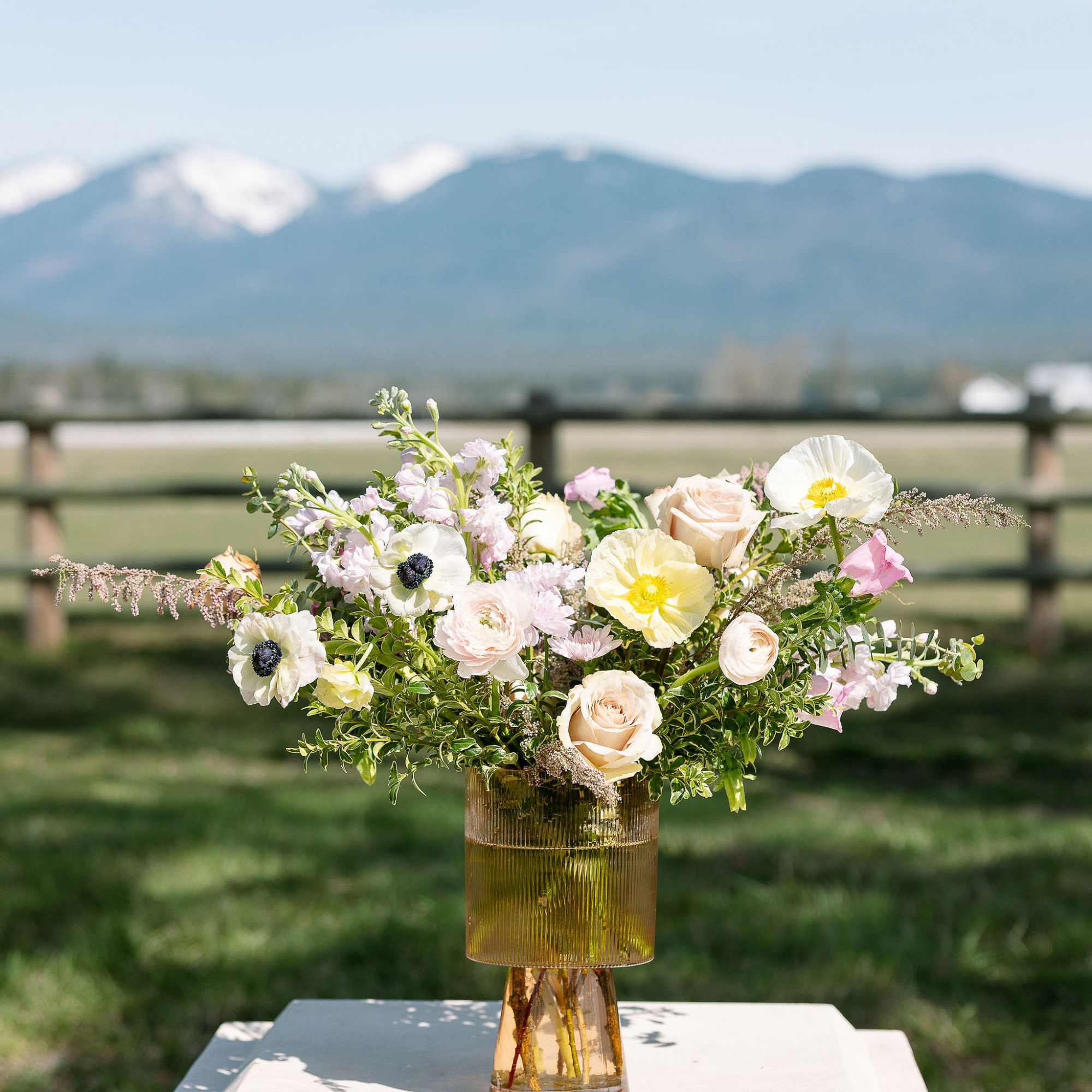 Pastel arrangement of roses, poppies, and anemones in an amber glass vase set outdoors.