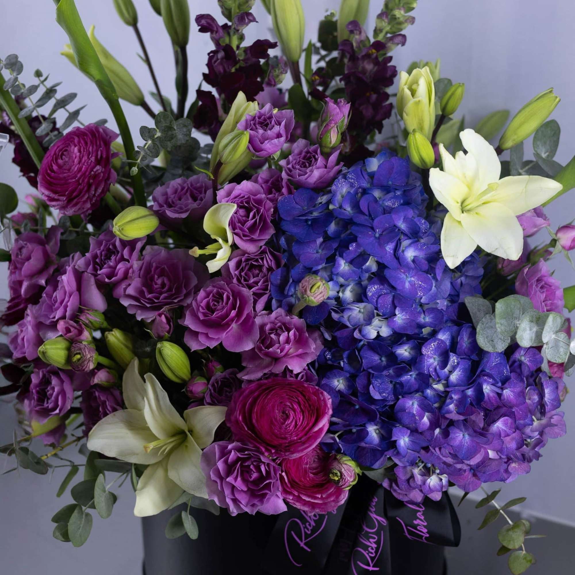 Purple and blue floral arrangement with white lilies in a black hat box