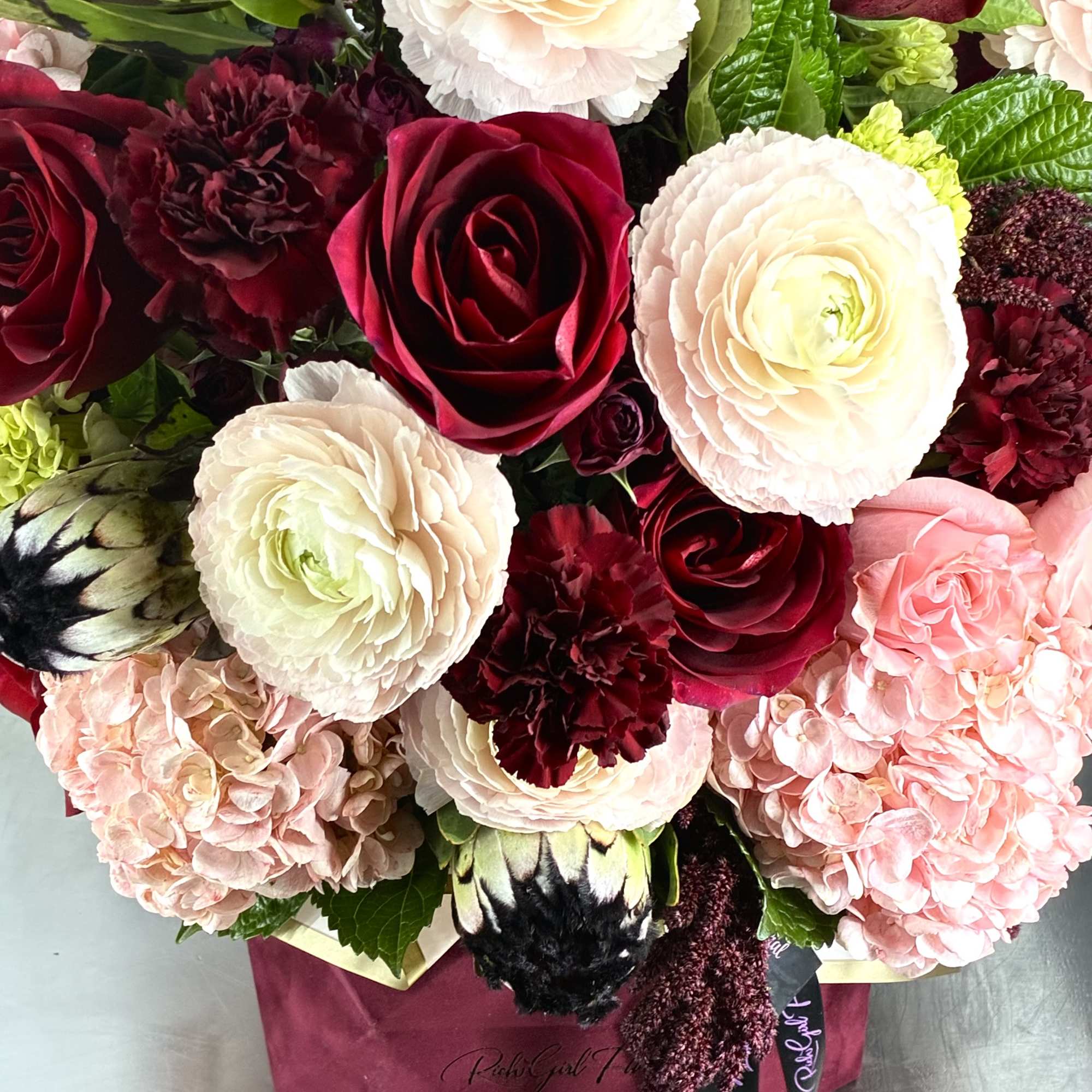 Bouquet of red roses, pale ranunculus, pink hydrangeas, and burgundy carnations in a box