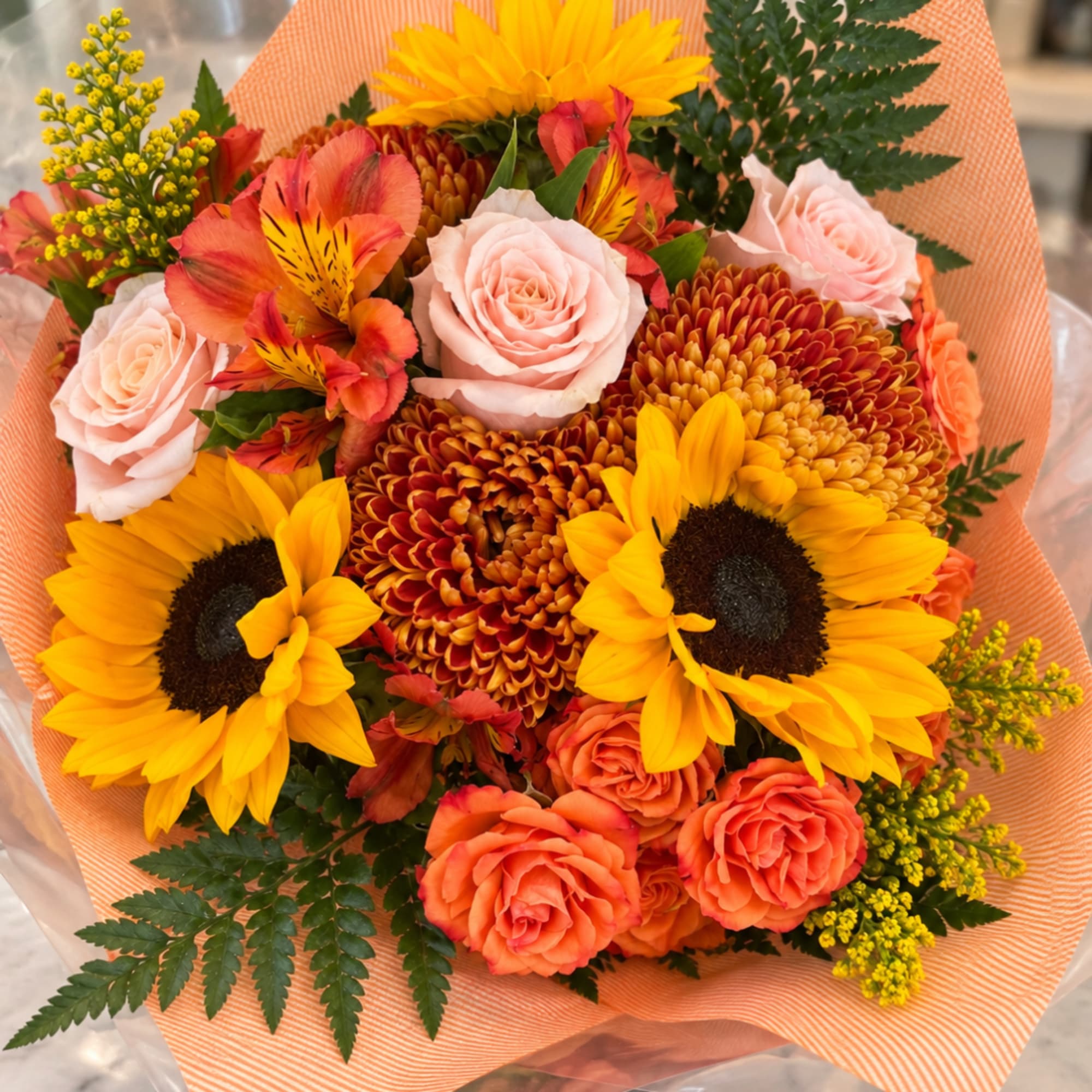 Mixed bouquet with sunflowers, roses, and orange blooms wrapped in peach paper