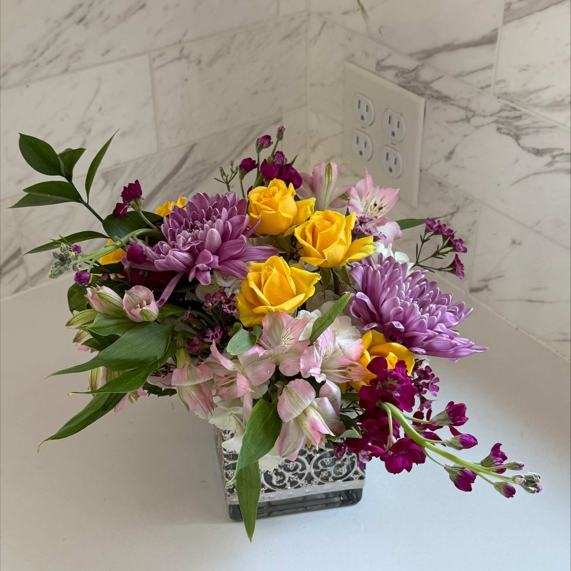 Mixed bouquet of yellow roses, purple mums, and pink alstroemeria in a square vase