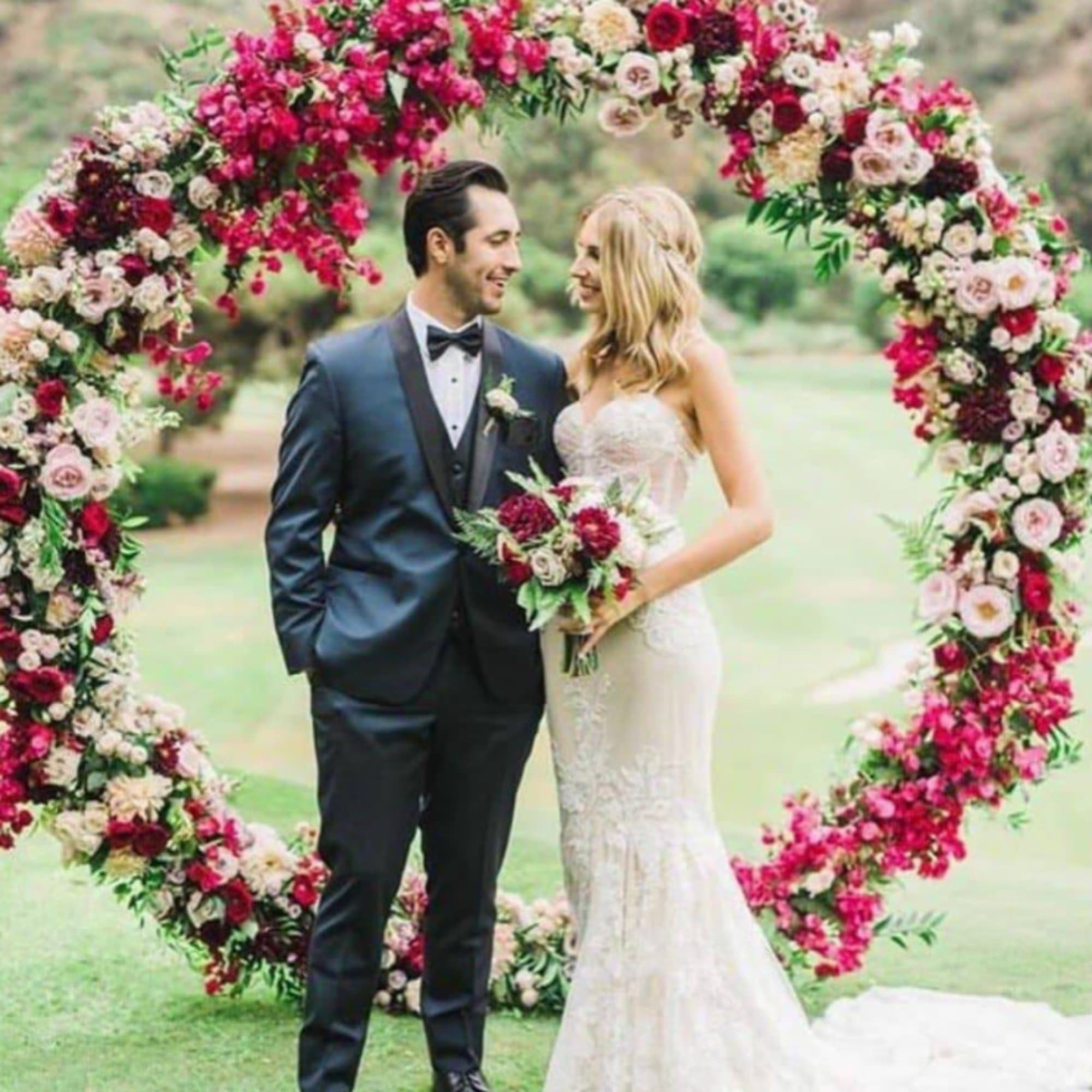 Bride and groom standing under a large floral arch at an outdoor wedding