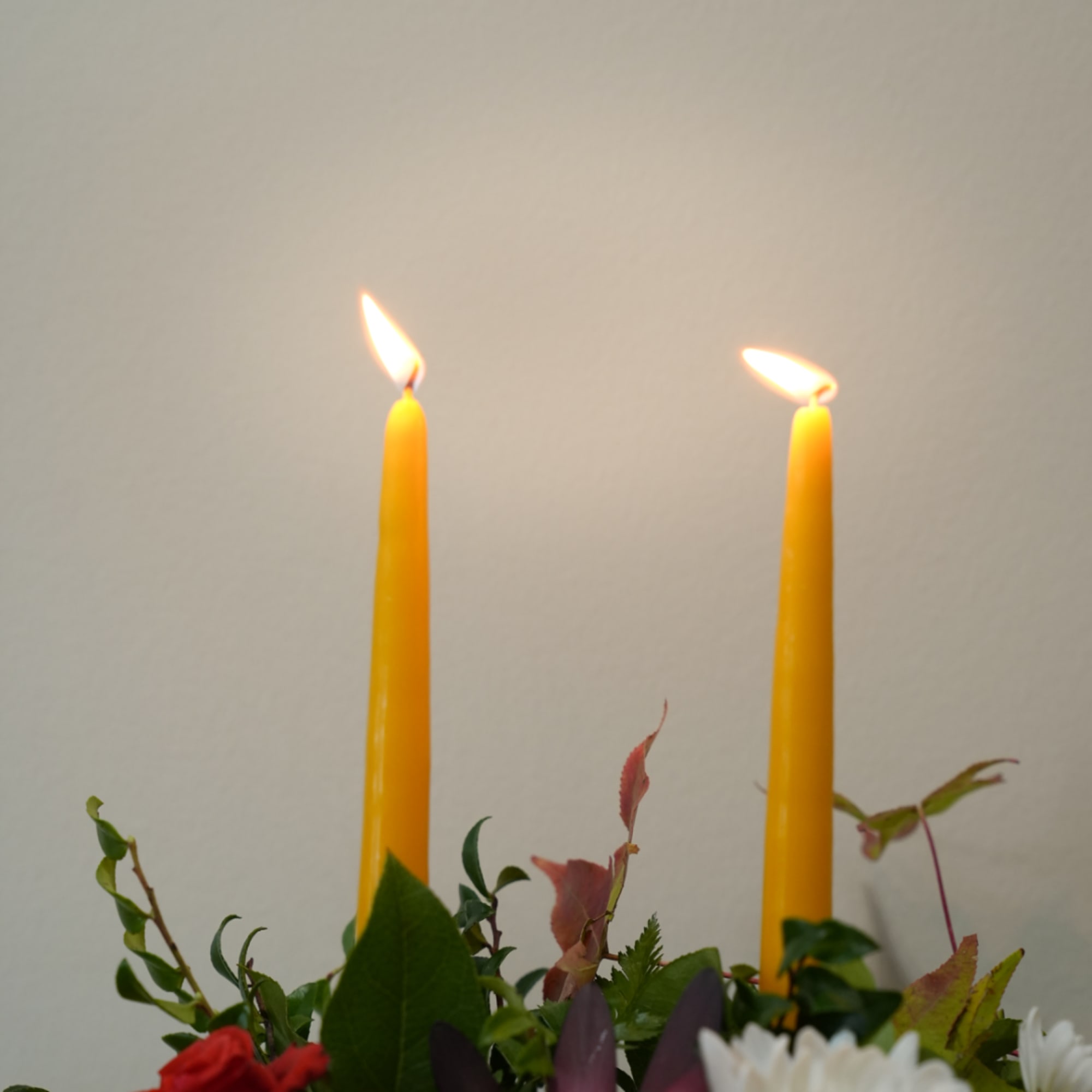 Two lit yellow taper candles above a floral arrangement with white daisies and greenery