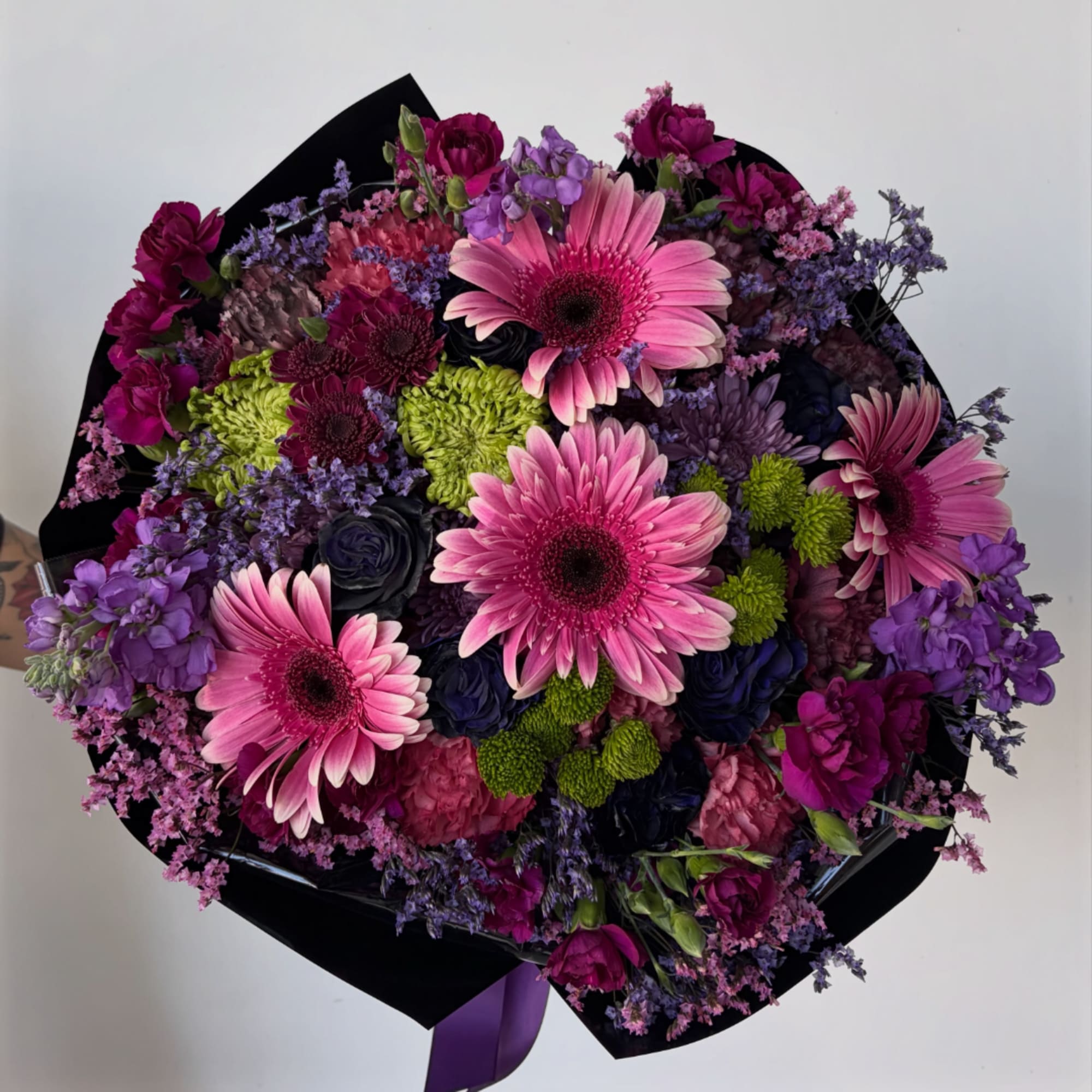 Bouquet of pink gerbera daisies, purple blooms, and green button mums in black wrap