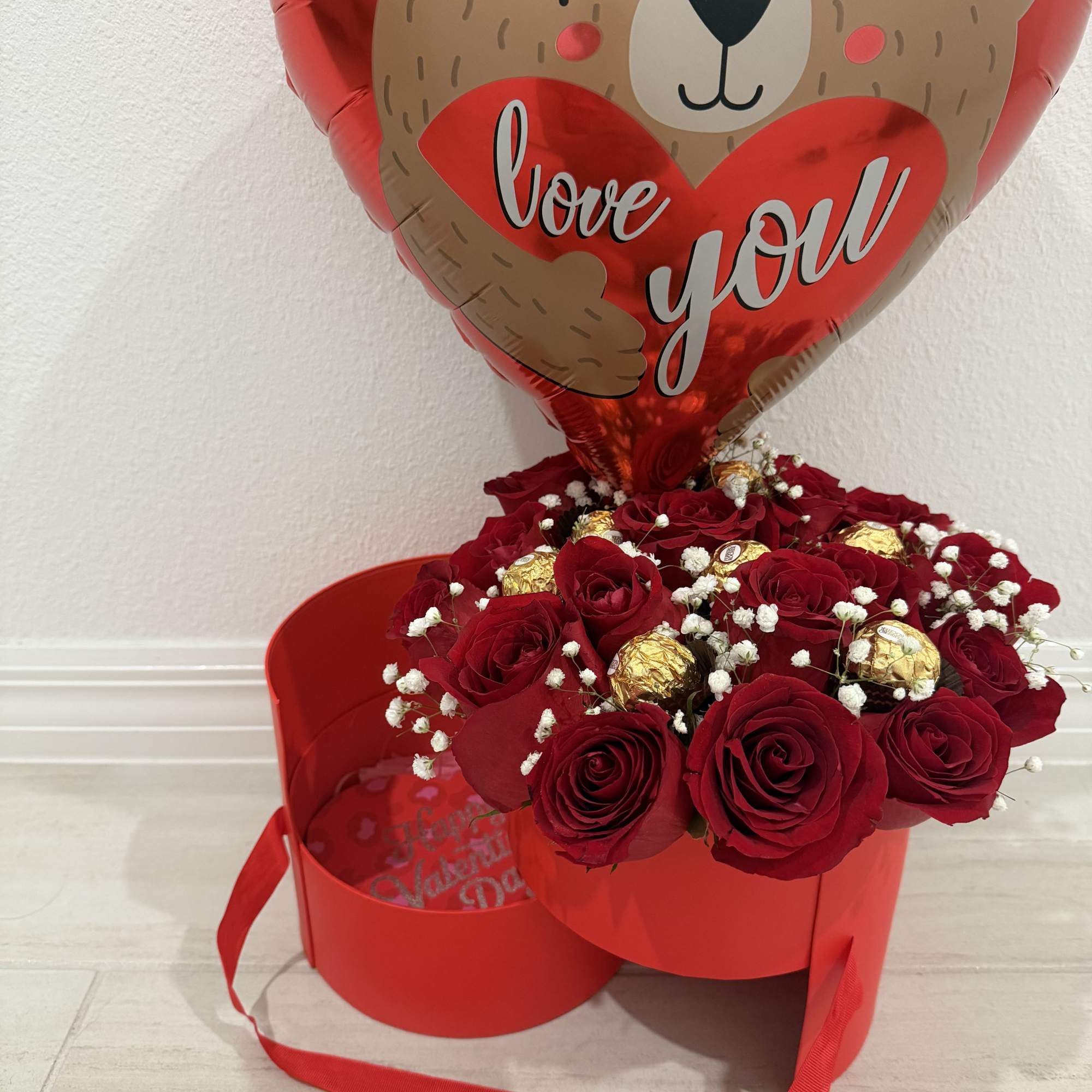 Red roses with baby’s breath and chocolates in a red box, topped by a bear balloon
