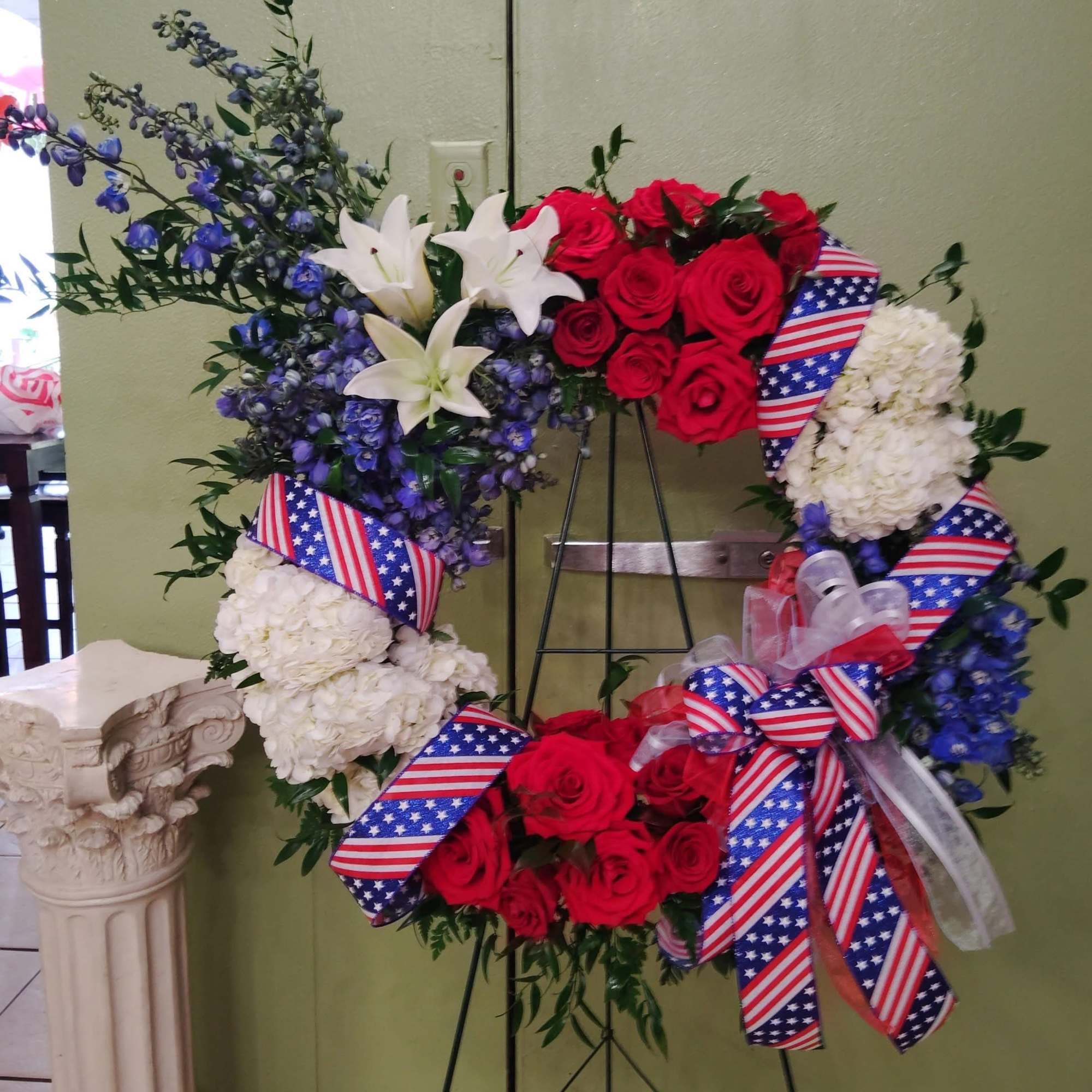 Floral wreath with red roses, white hydrangeas, blue flowers, and patriotic ribbon