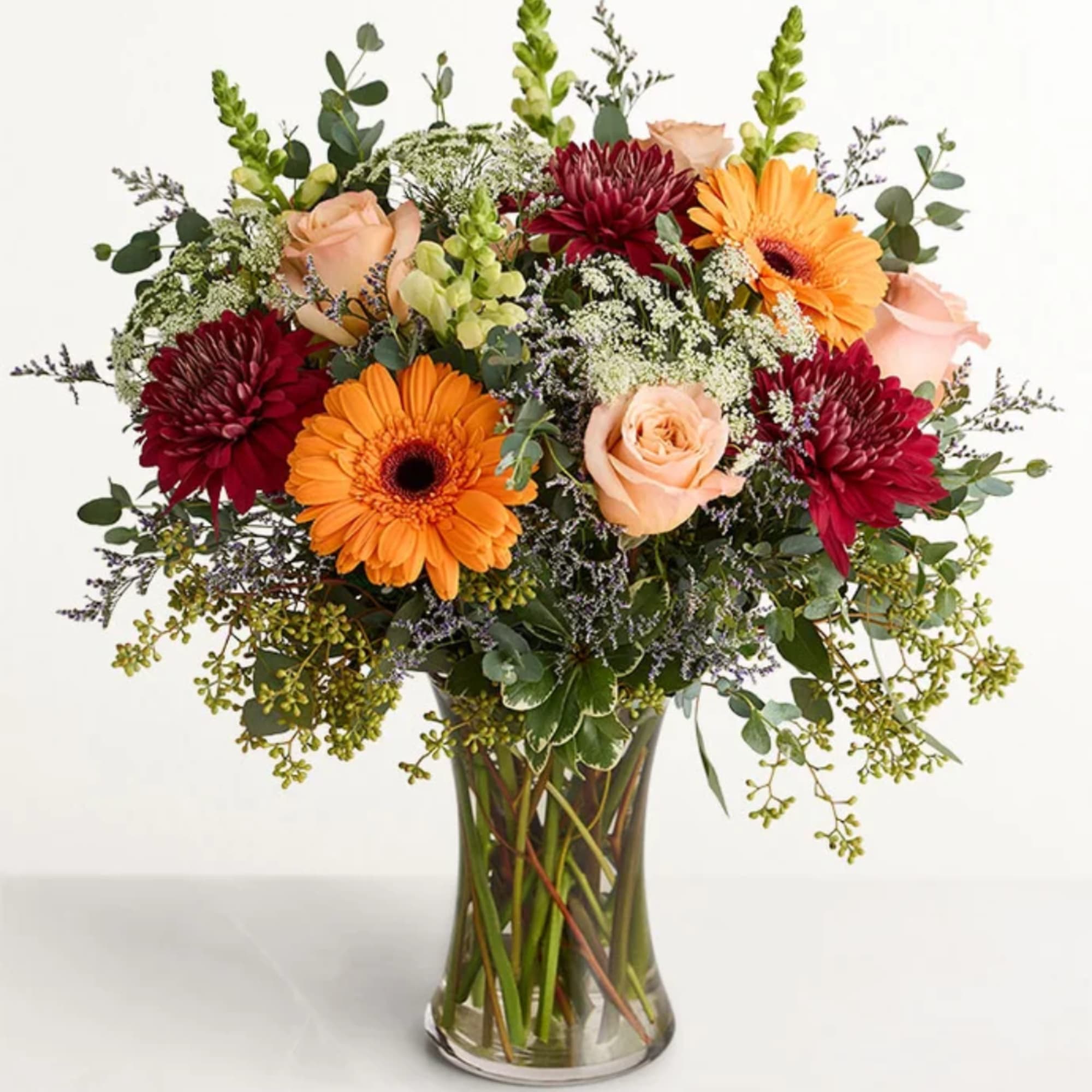 Mixed bouquet of orange gerberas, burgundy mums, and peach roses in a glass vase