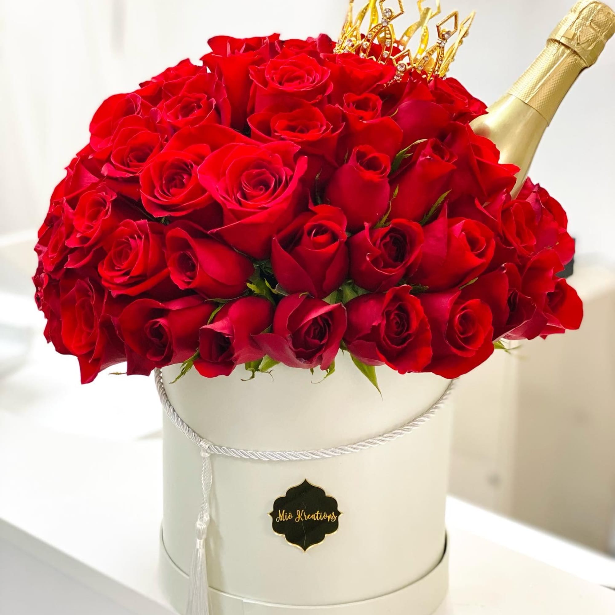Red roses arranged in a white hat box with a gold bottle topper