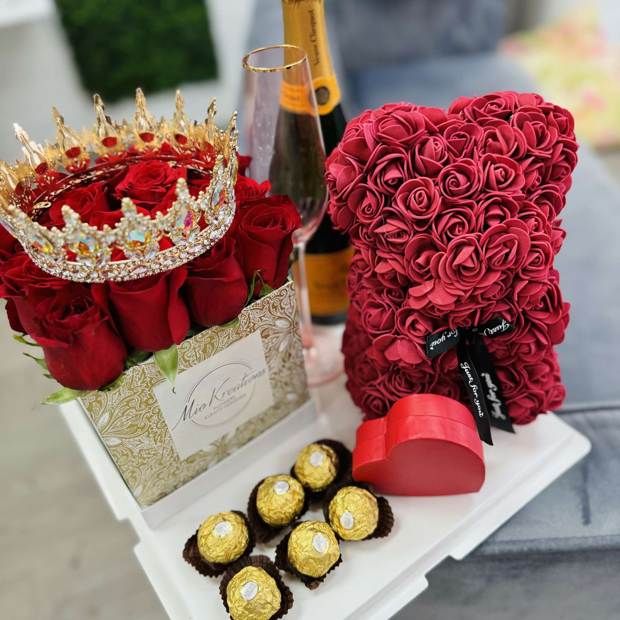 Red rose arrangement with a jeweled crown, champagne bottle, heart box, and chocolates
