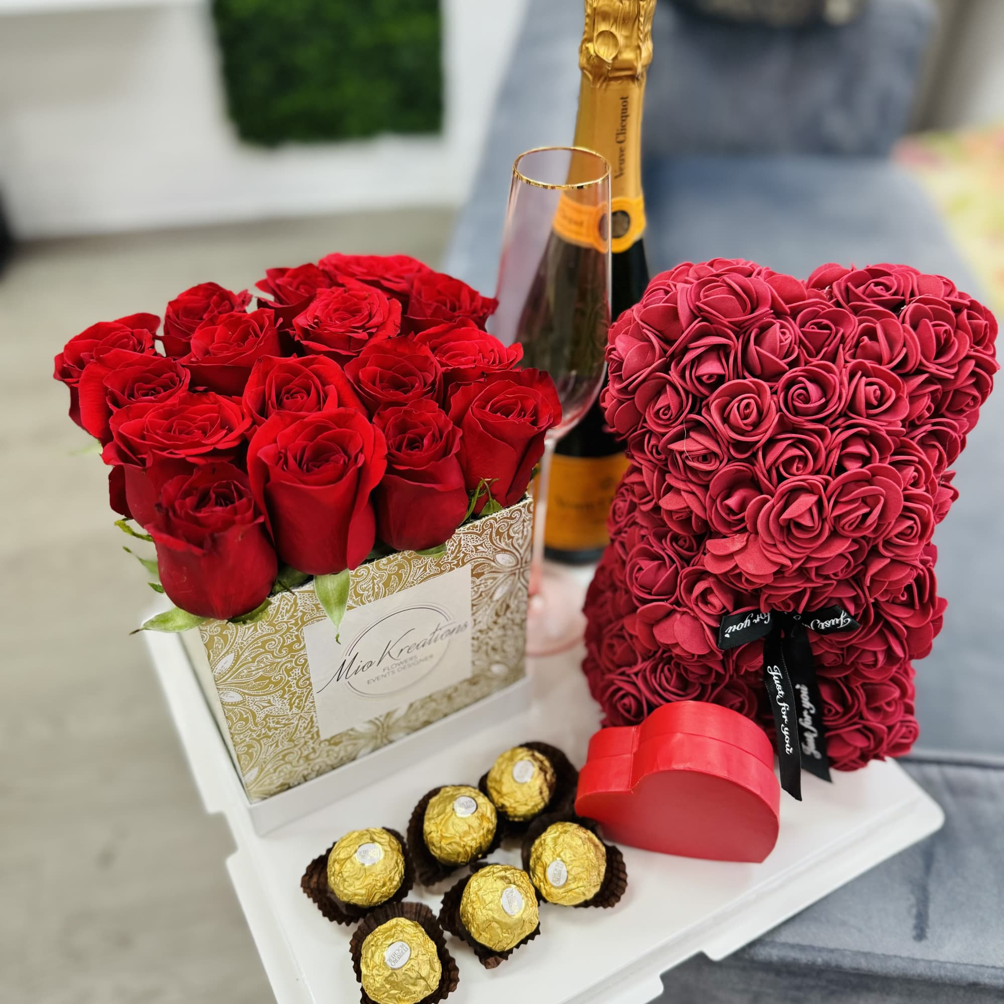 Red roses in a decorative box with a champagne bottle, glass, heart box, and chocolates.