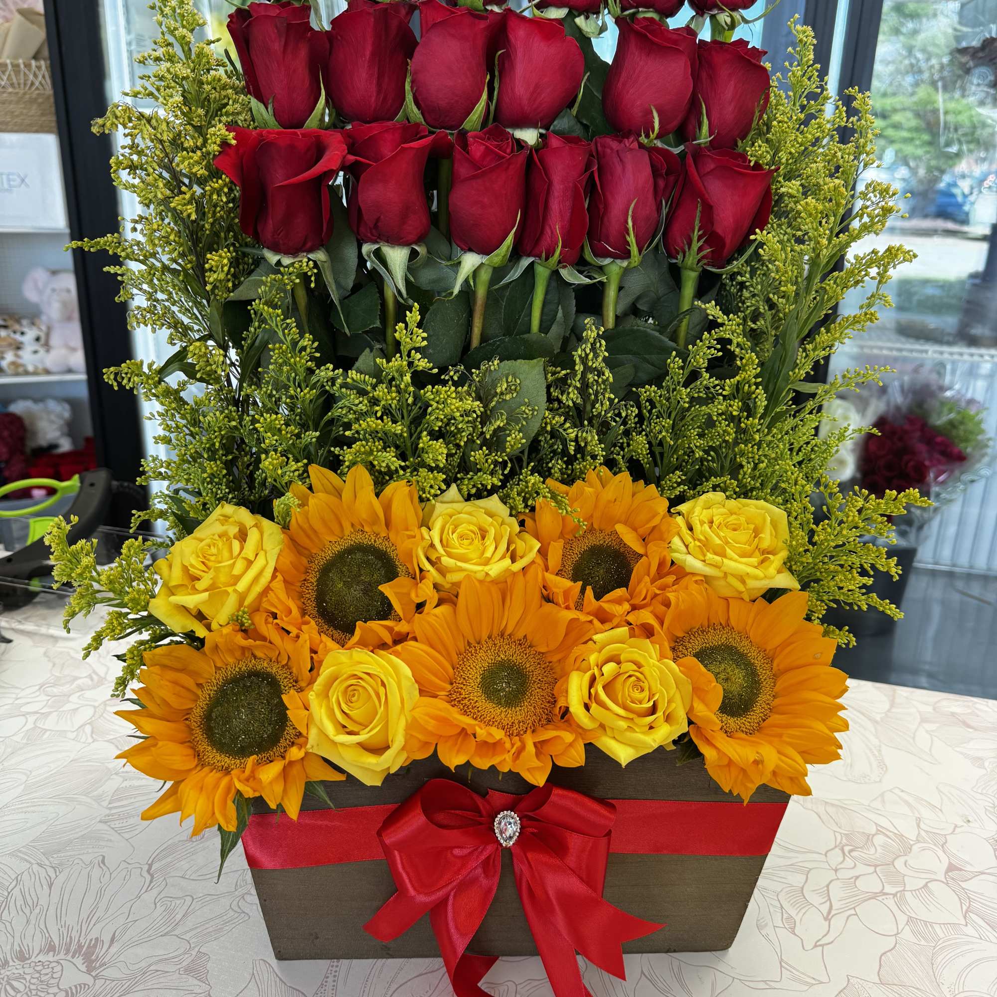 Tiered arrangement of red roses, yellow roses, and sunflowers in a box with a red bow