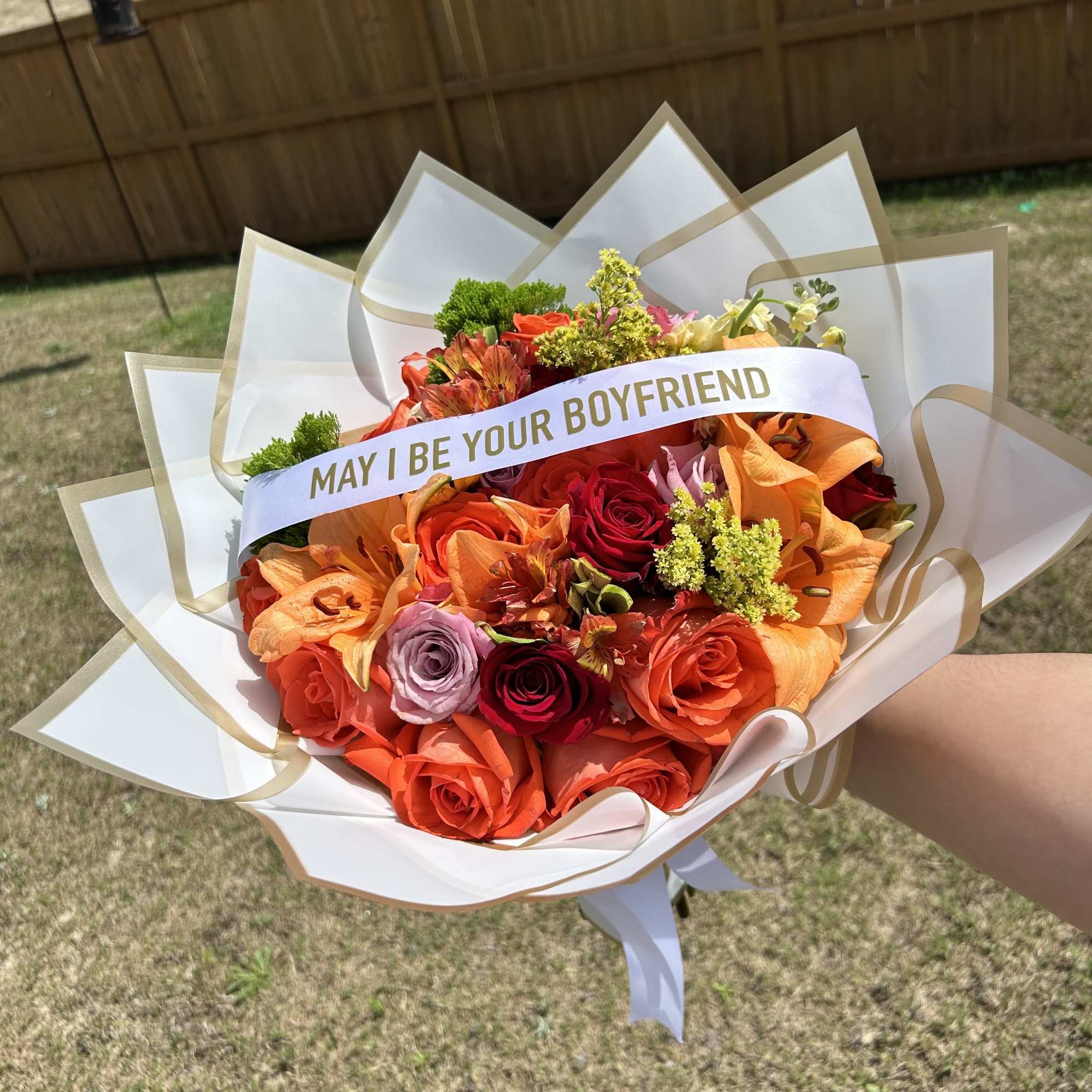 Bouquet of orange and red roses with lilies and a ribbon reading "MAY I BE YOUR BOYFRIEND"