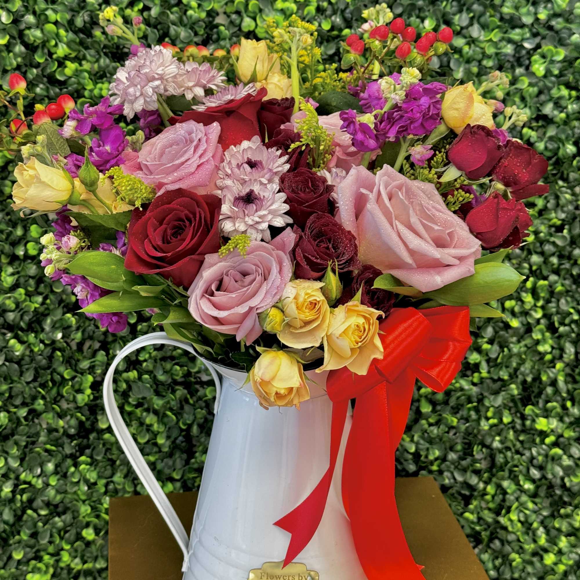 Mixed bouquet of roses and small blooms in a white metal pitcher with a red ribbon