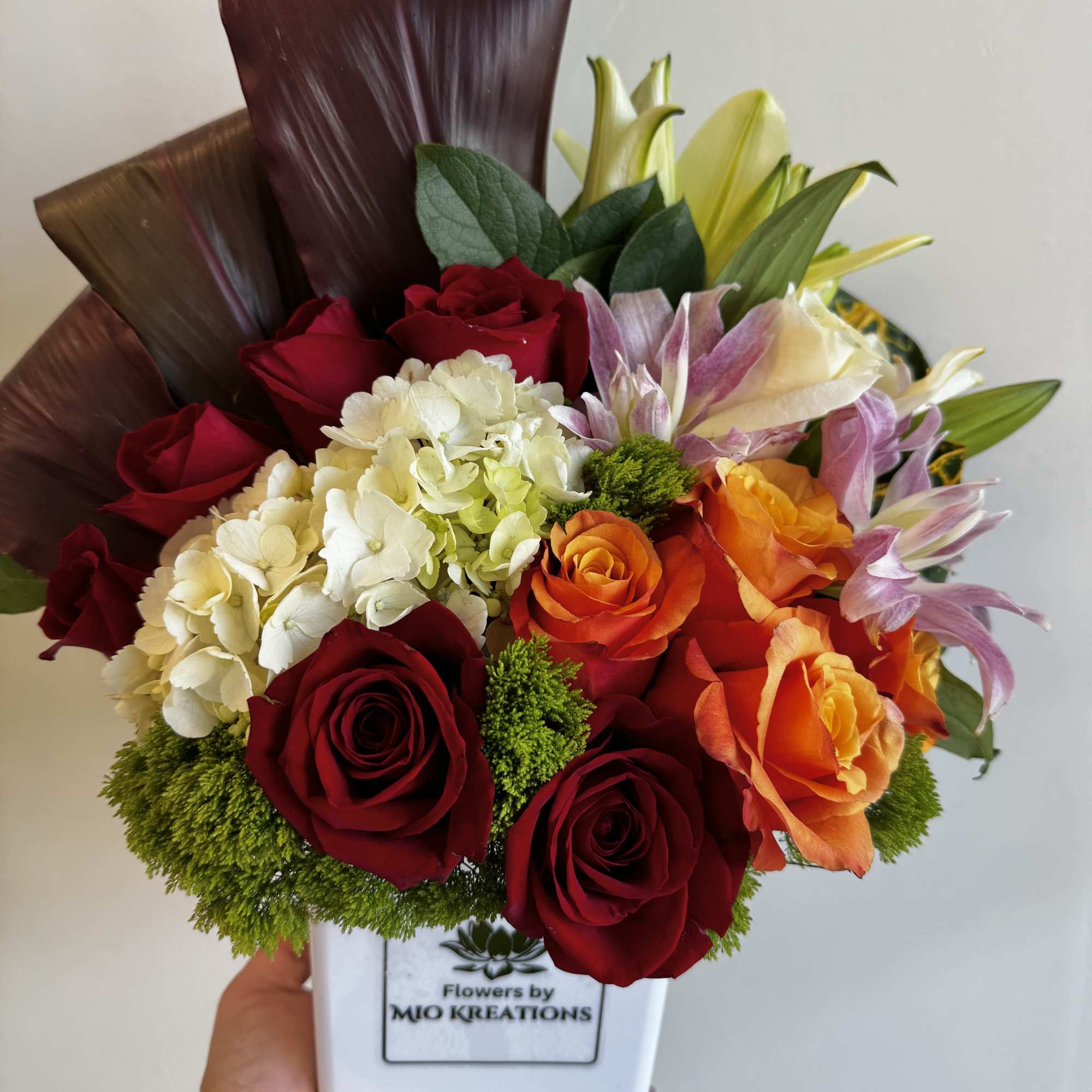 Mixed bouquet of red roses, hydrangea, lilies, and orange blooms in a white vase