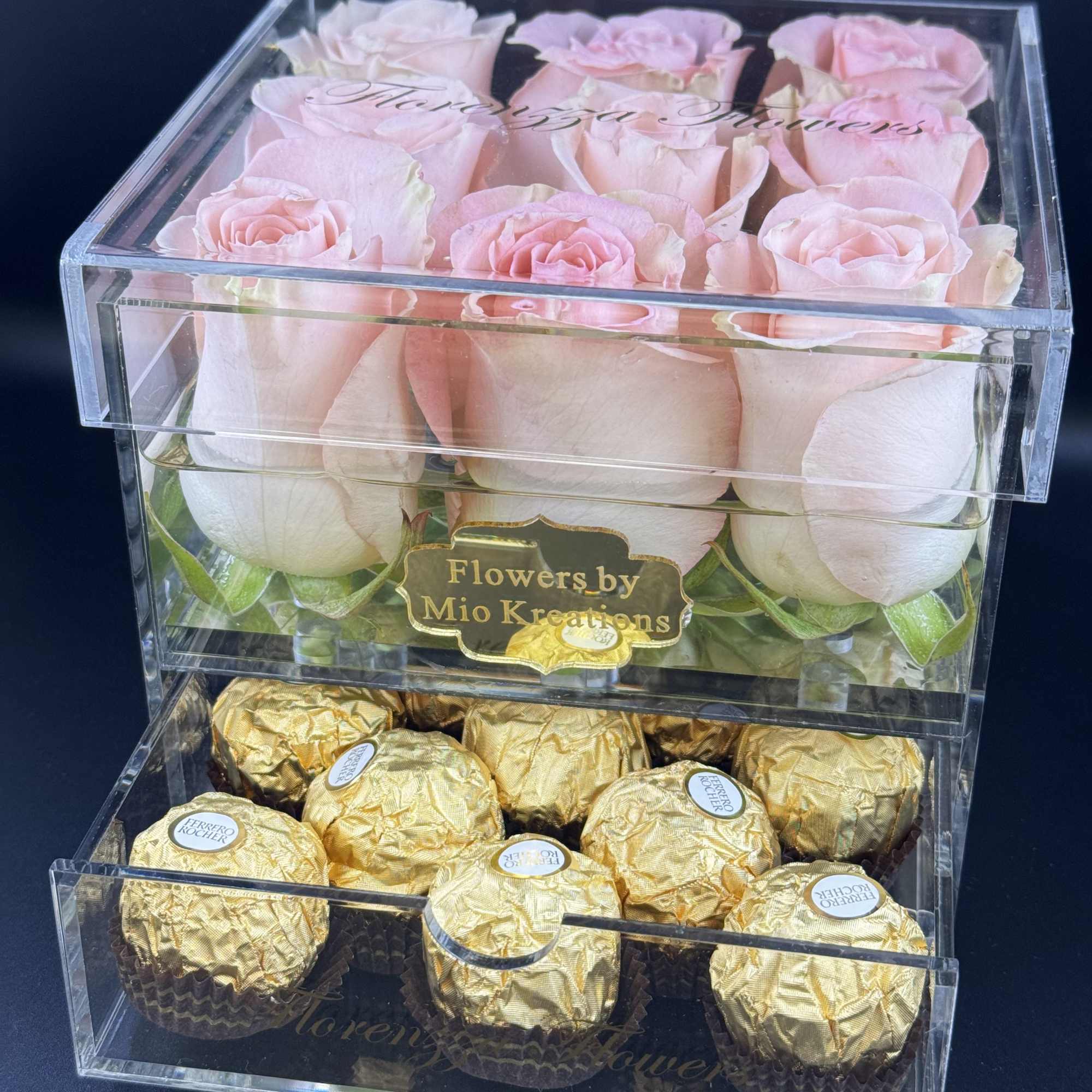 Pink roses in a clear box above gold-wrapped chocolates.