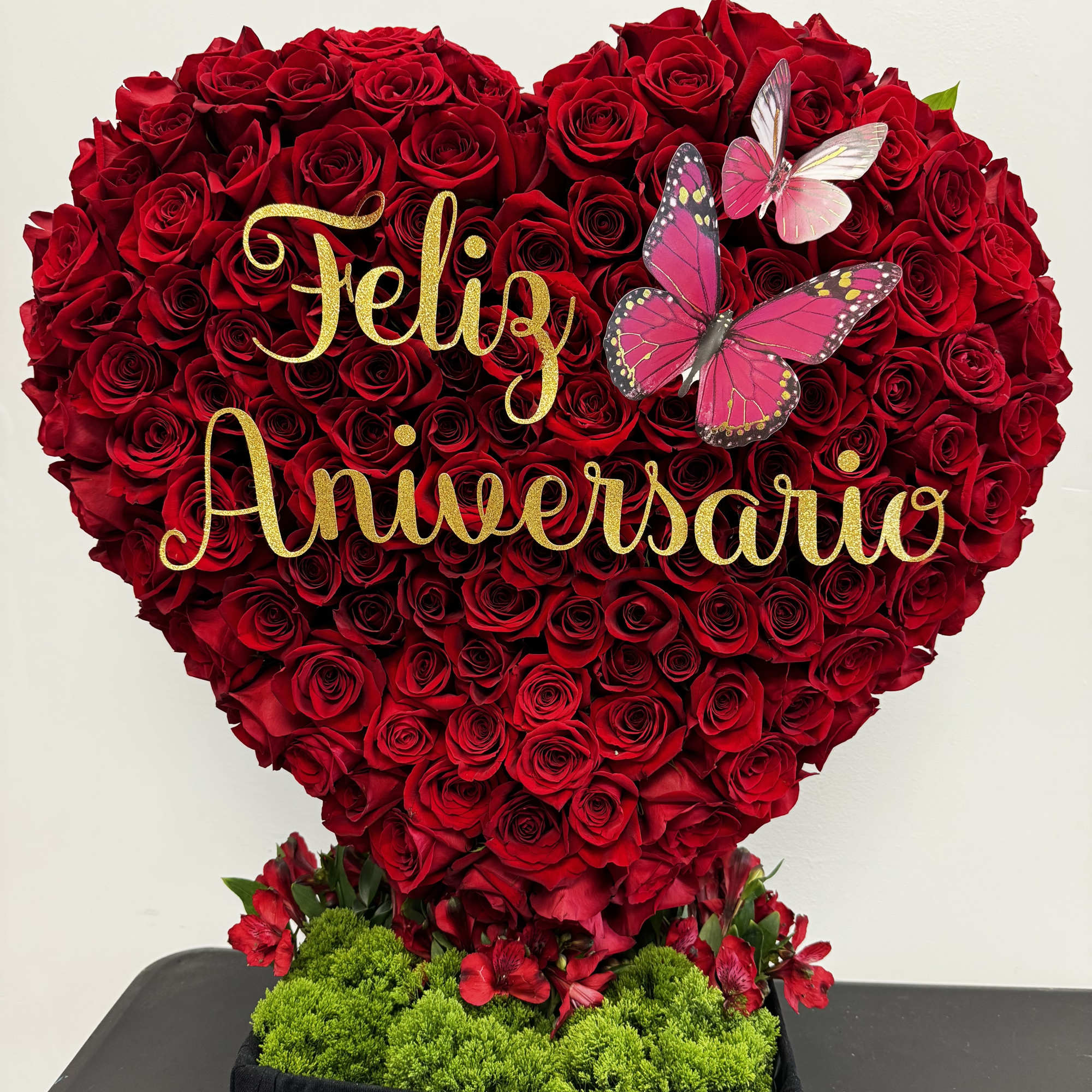 Heart-shaped red rose arrangement with gold "Feliz Aniversario" lettering and butterflies