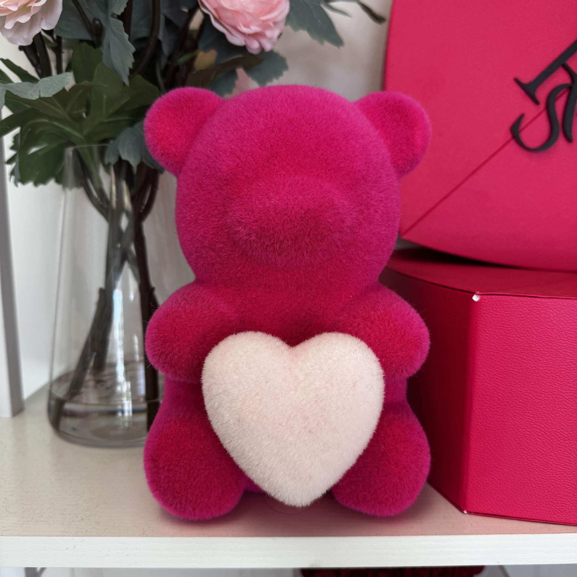 Pink teddy bear with a white heart beside pink flowers in a vase