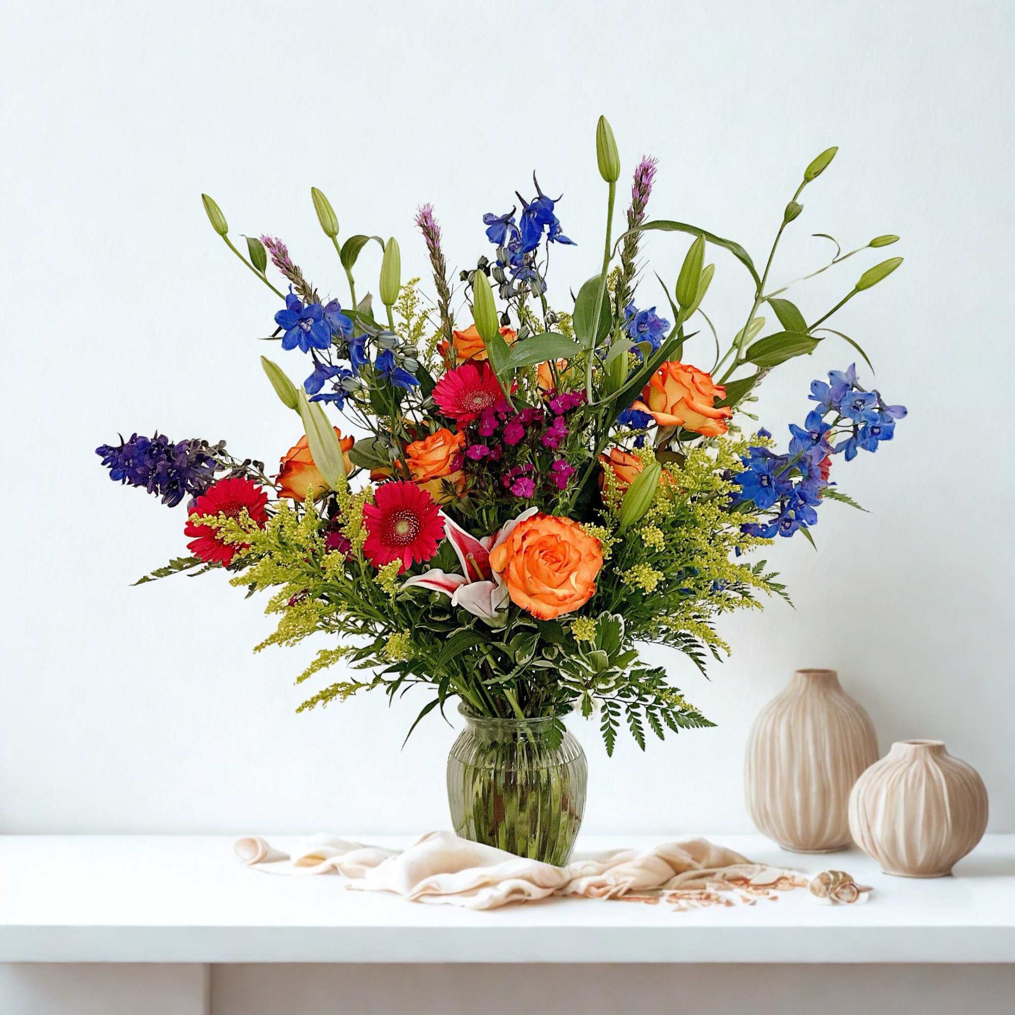 Mixed bouquet of orange roses, blue delphinium, and red gerbera daisies in a glass vase