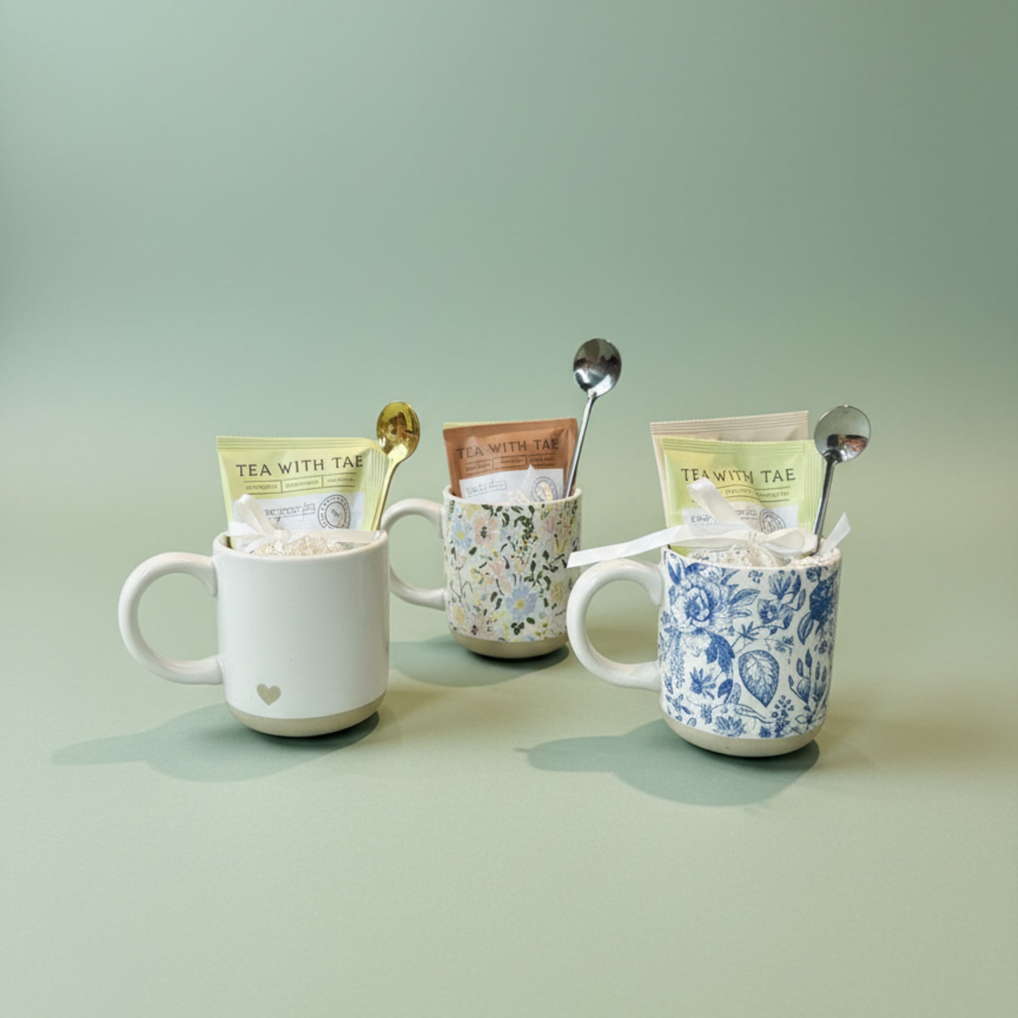 Three tea gift mugs with packets and spoons on a pale green background