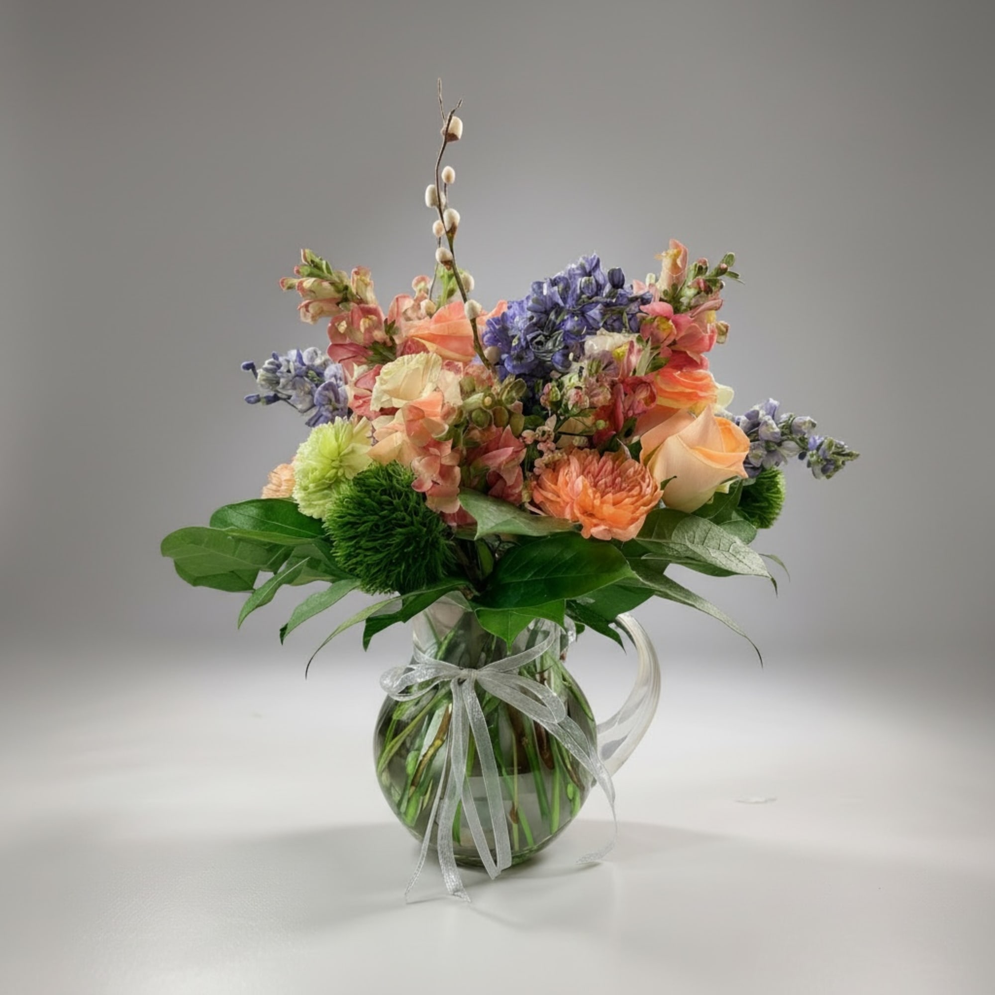 Mixed pastel bouquet in a clear glass pitcher vase with a ribbon