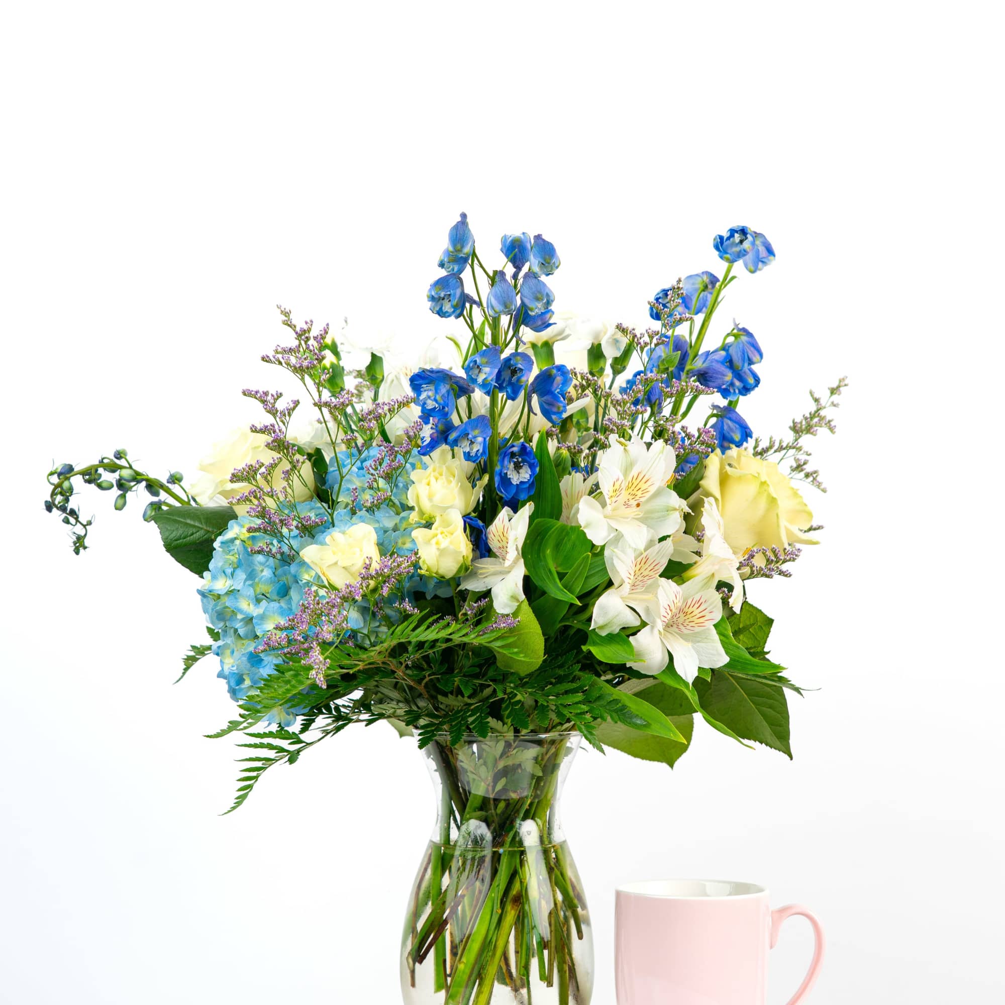 Tall blue and white mixed flower arrangement in a clear vase beside a pink mug