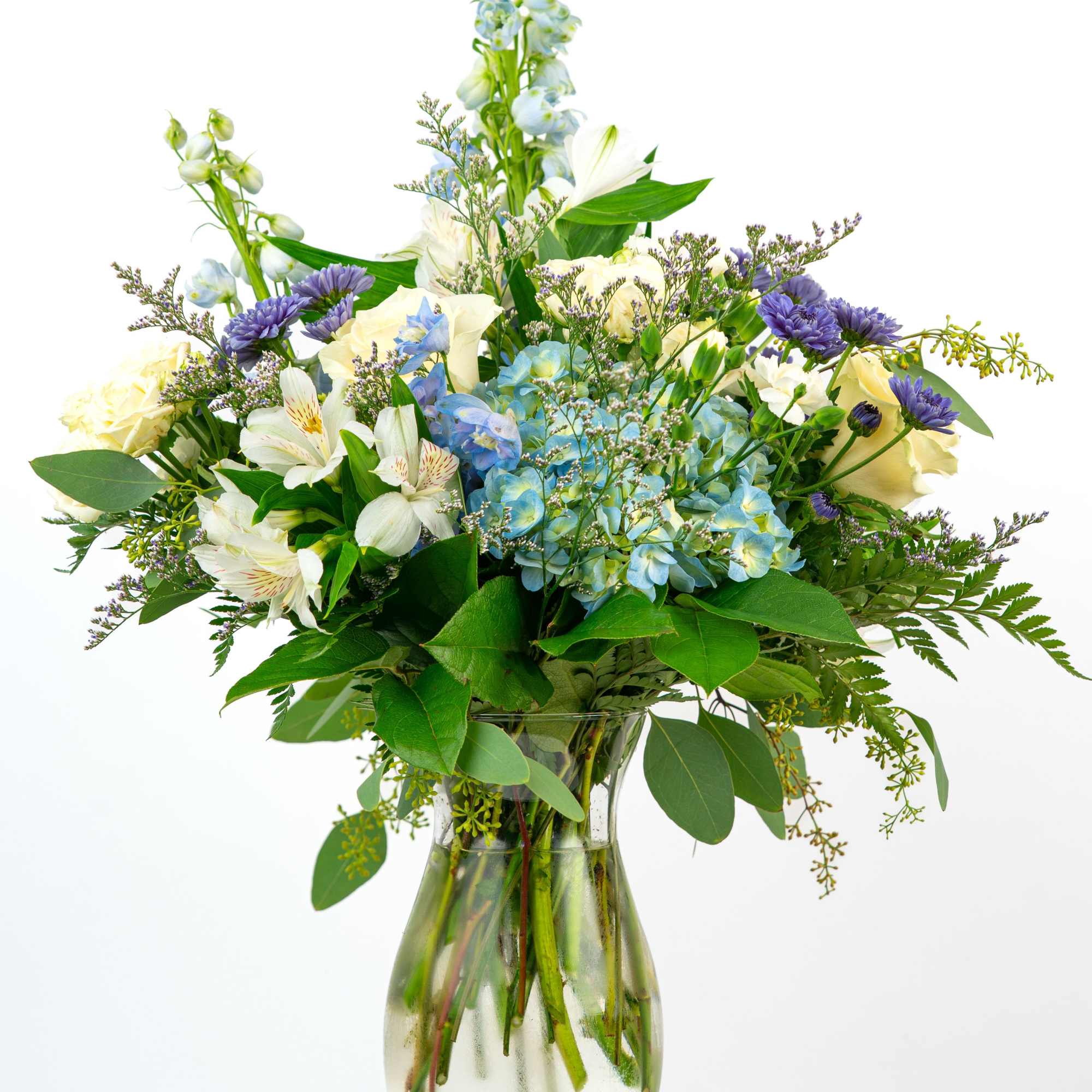 Tall mixed bouquet of white, cream, blue, and purple flowers in a clear glass vase
